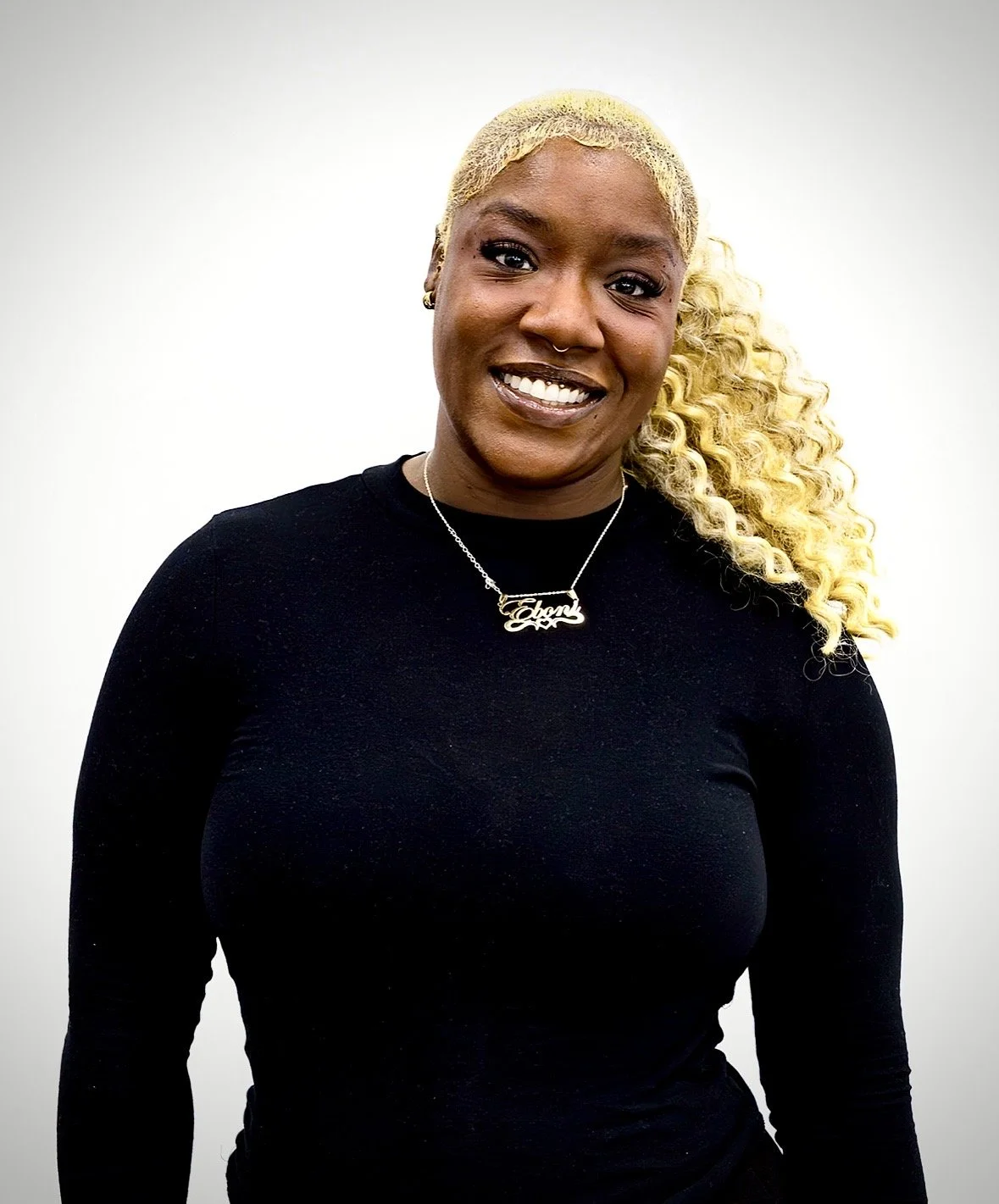 A woman with blonde, curly hair in a ponytail, wearing a black top and a name necklace, smiling against a plain white background.