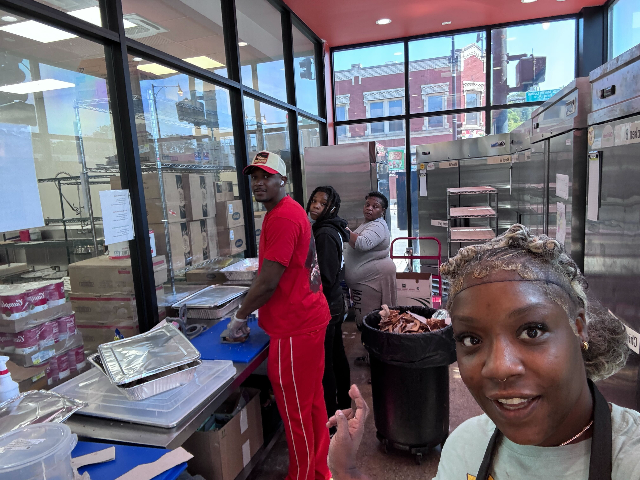 A woman taking a selfie in a busy commercial kitchen with three people preparing food. The background shows stainless steel equipment and large windows, with two young men and one woman working behind her.
