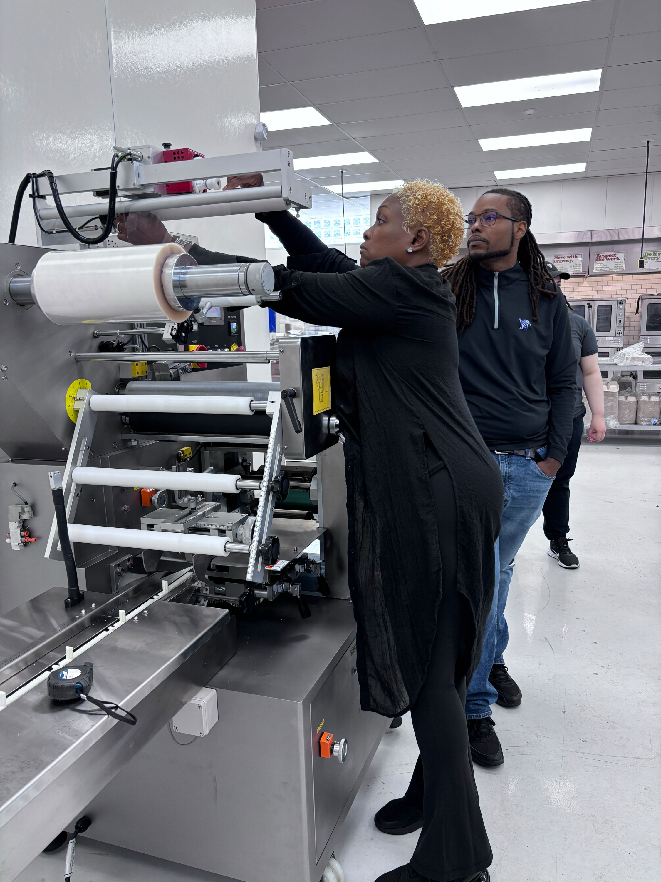 Two people observing a woman operating a large industrial machine in a factory or laboratory setting.