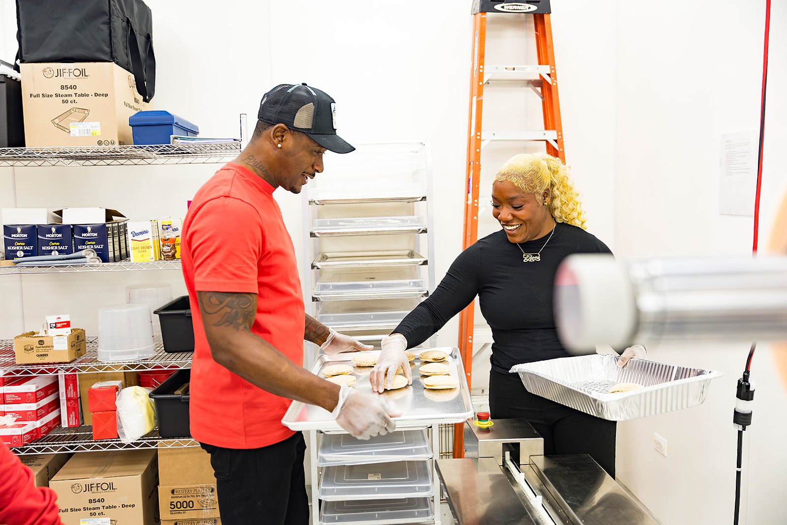 Two people preparing sandwiches in a food kitchen or storage room. One person is wearing a red shirt and black hat, and the other is wearing a black shirt with blonde curly hair. They are working with trays of bread and ingredients.