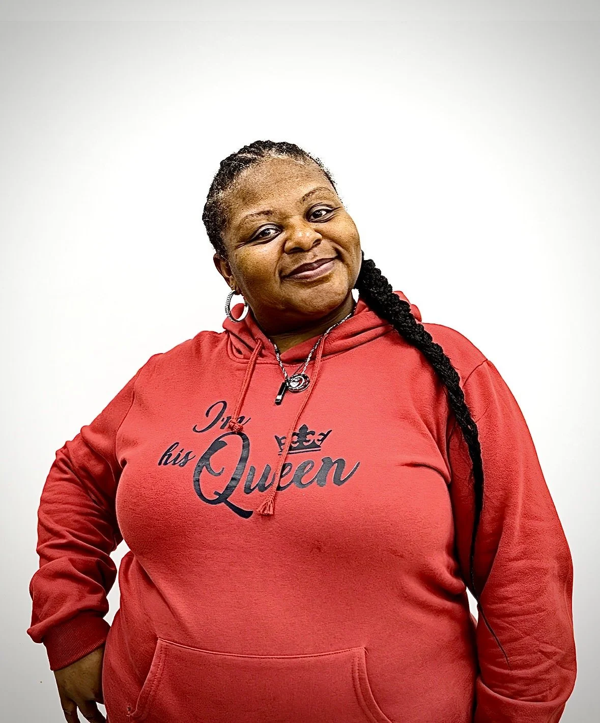 Woman wearing a red hoodie with the text "I'm his Queen" smiling at the camera, with her hair in braids, standing against a plain white background.