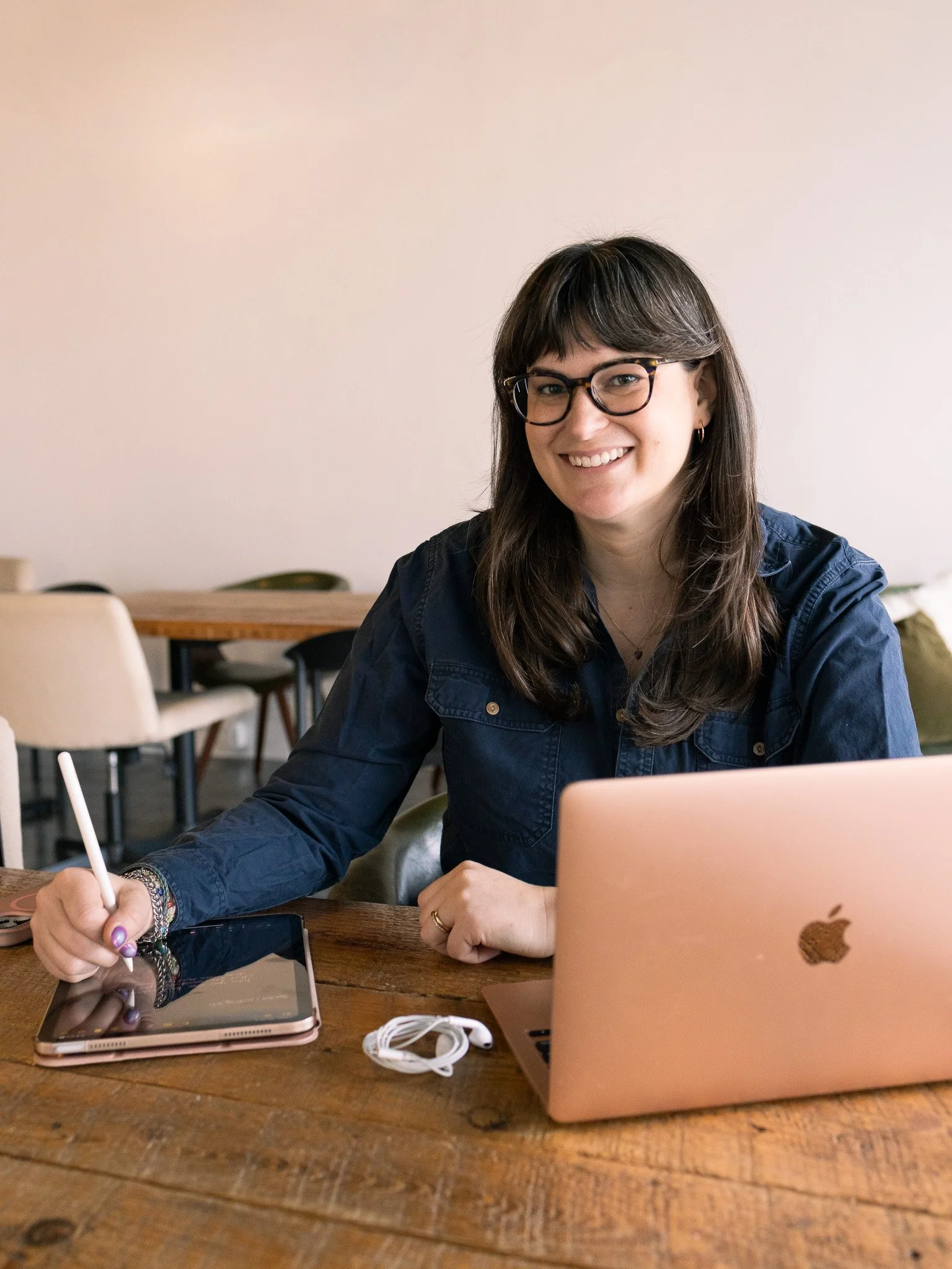 Nicole Parker sitting at a table with her laptop open beside her, she is holding a pen and writing on an iPad, while she looks forward and smiles