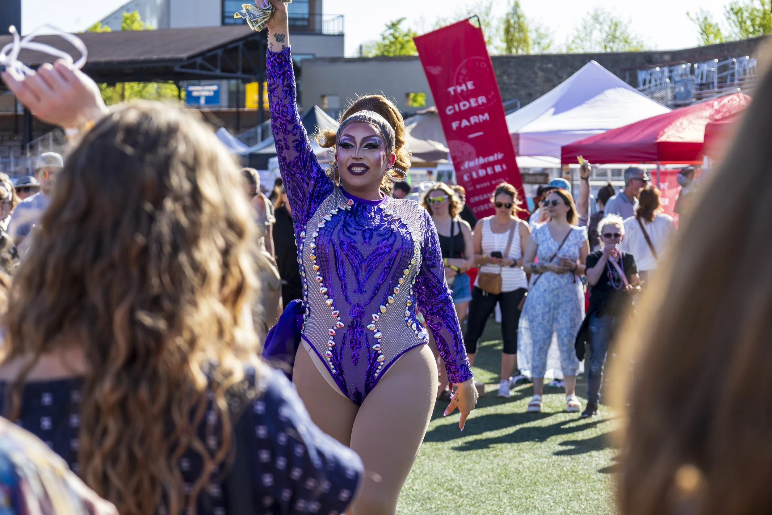 A drag performer in a purple, sequined costume waving to an audience at Uncork Me, with wine tents and a crowd in the background.