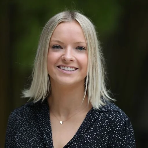 A young woman with blonde hair smiling outdoors, wearing a dark, polka-dot top and a delicate necklace.