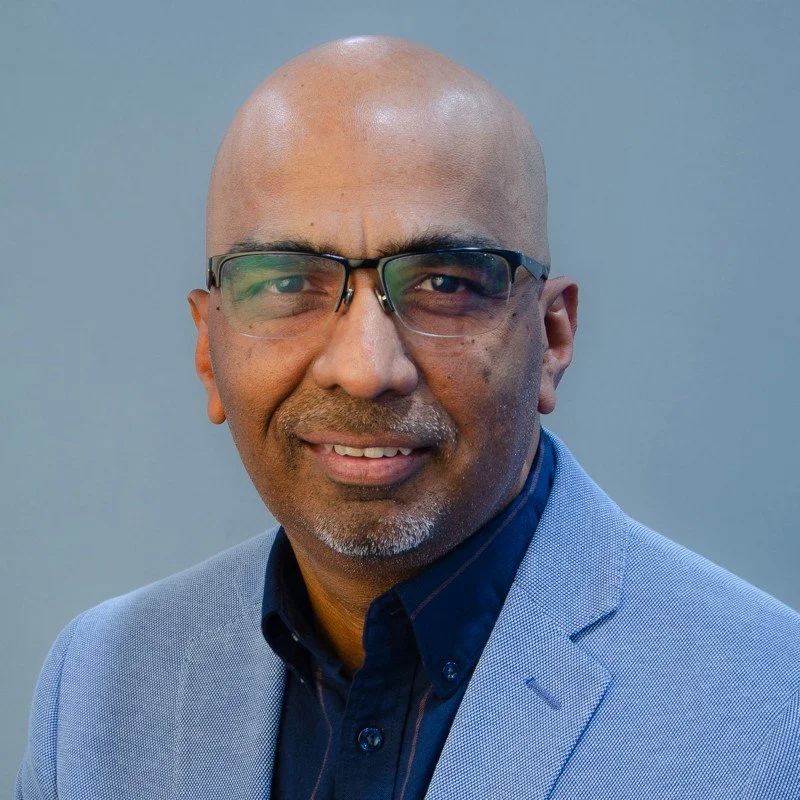 A middle-aged man with a shaved head wearing glasses, a light blue blazer, and a dark button-up shirt smiling against a plain blue background.
