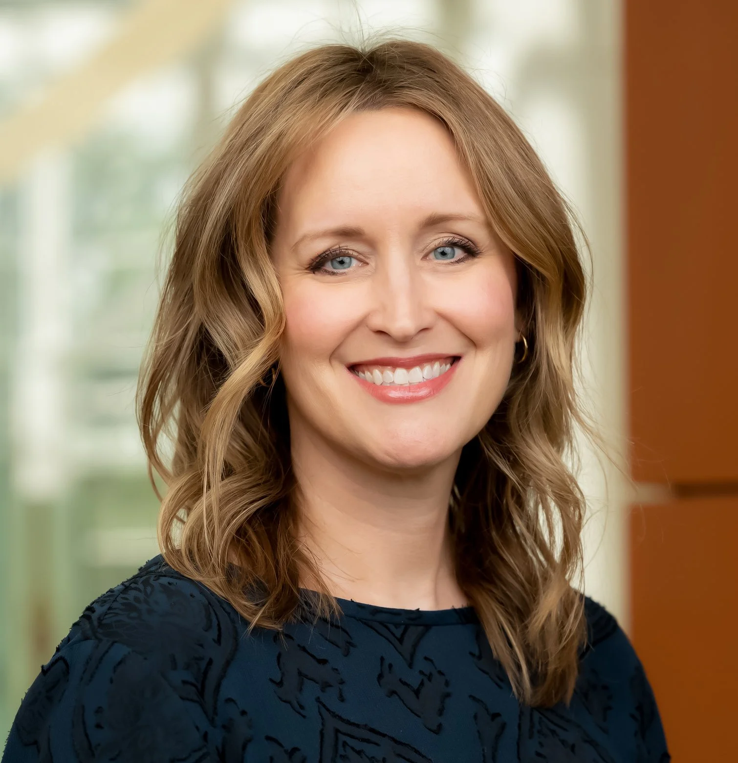 A smiling woman with light brown hair in front of a window background