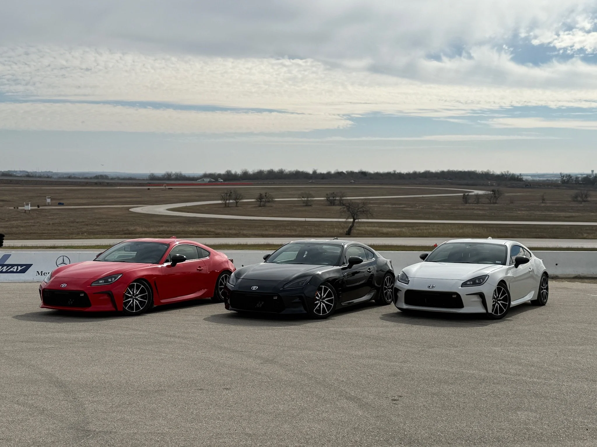 Three Toyota GR86's in red, black, and white, track prepped and ready. parked side by side on a race track with an open landscape and cloudy sky in the background.