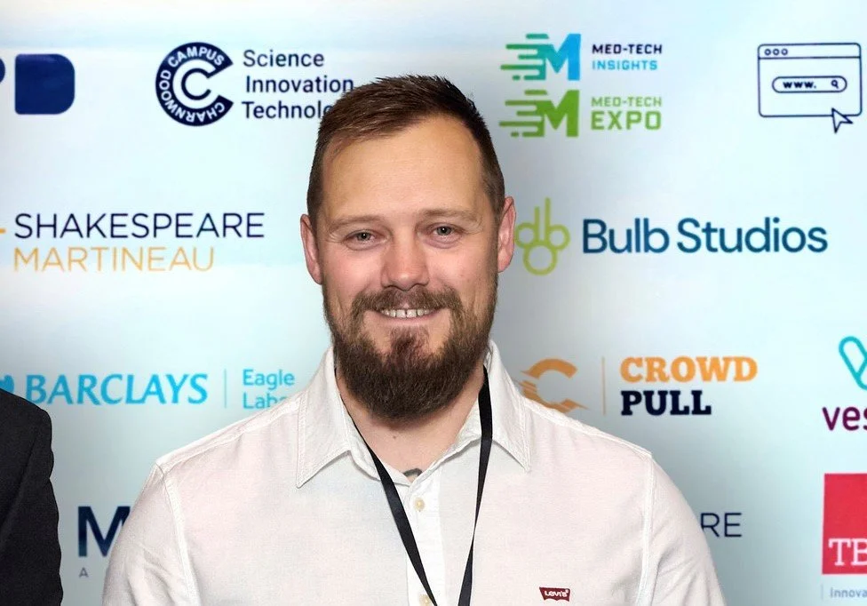 A man with dark hair and a beard in a white shirt, standing in front of a banner with lots of logos on it