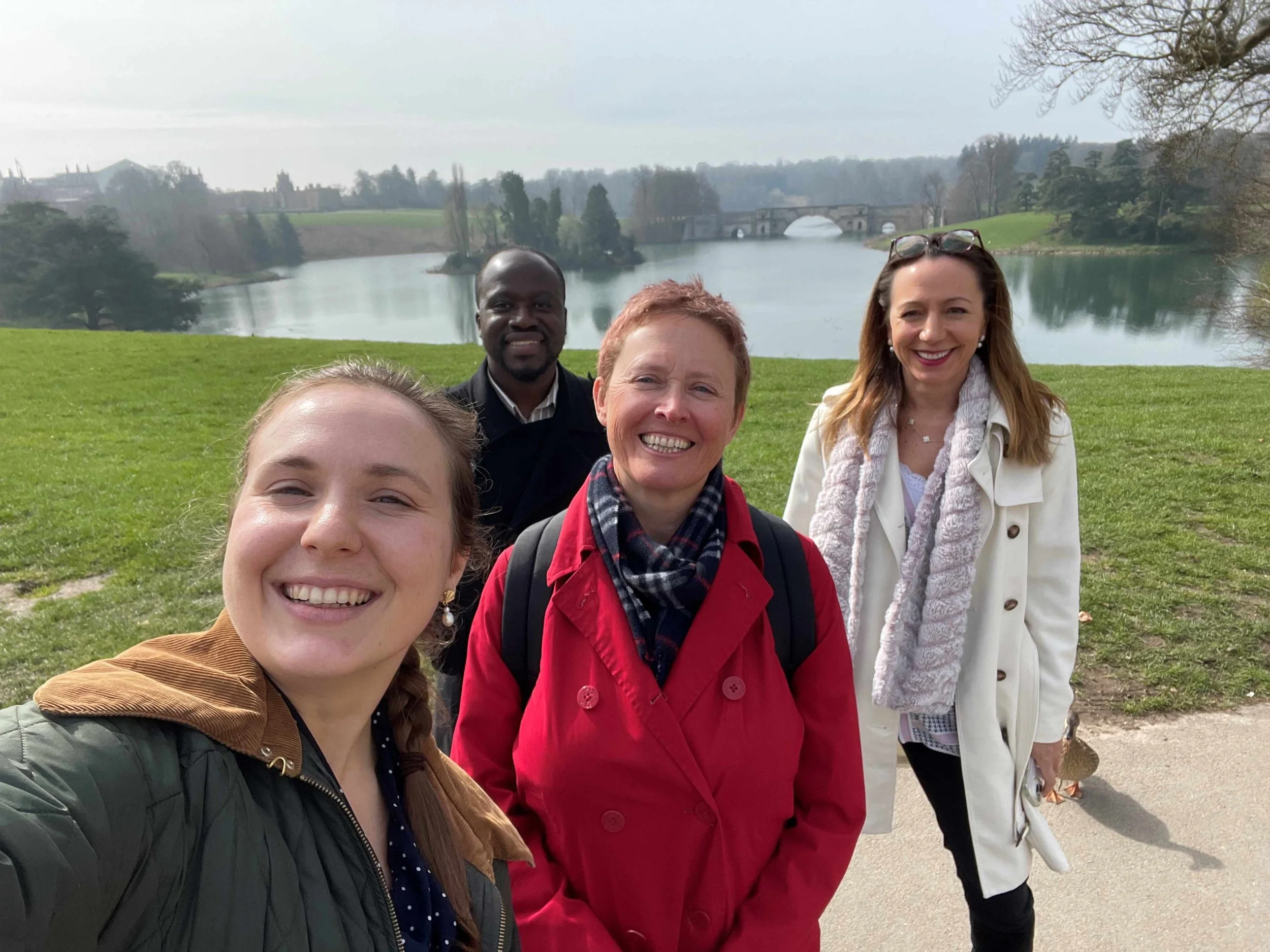 Four people (three women and one man) all smiling, standing in front of a lake, with a bridge in the background. All the people are smiling