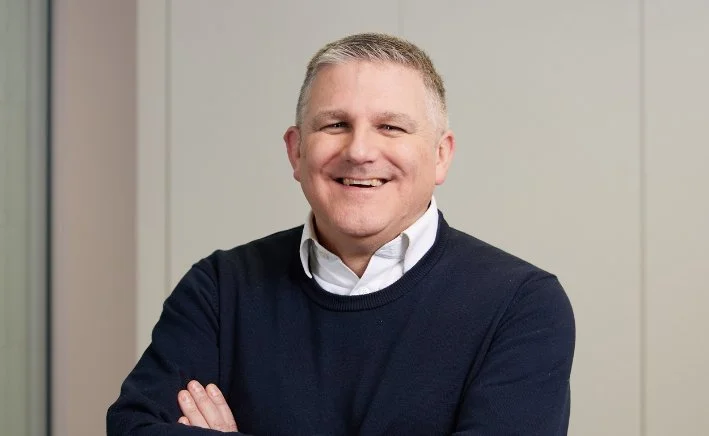 A man in a blue jumper and white short with his arms folded. He is smiling