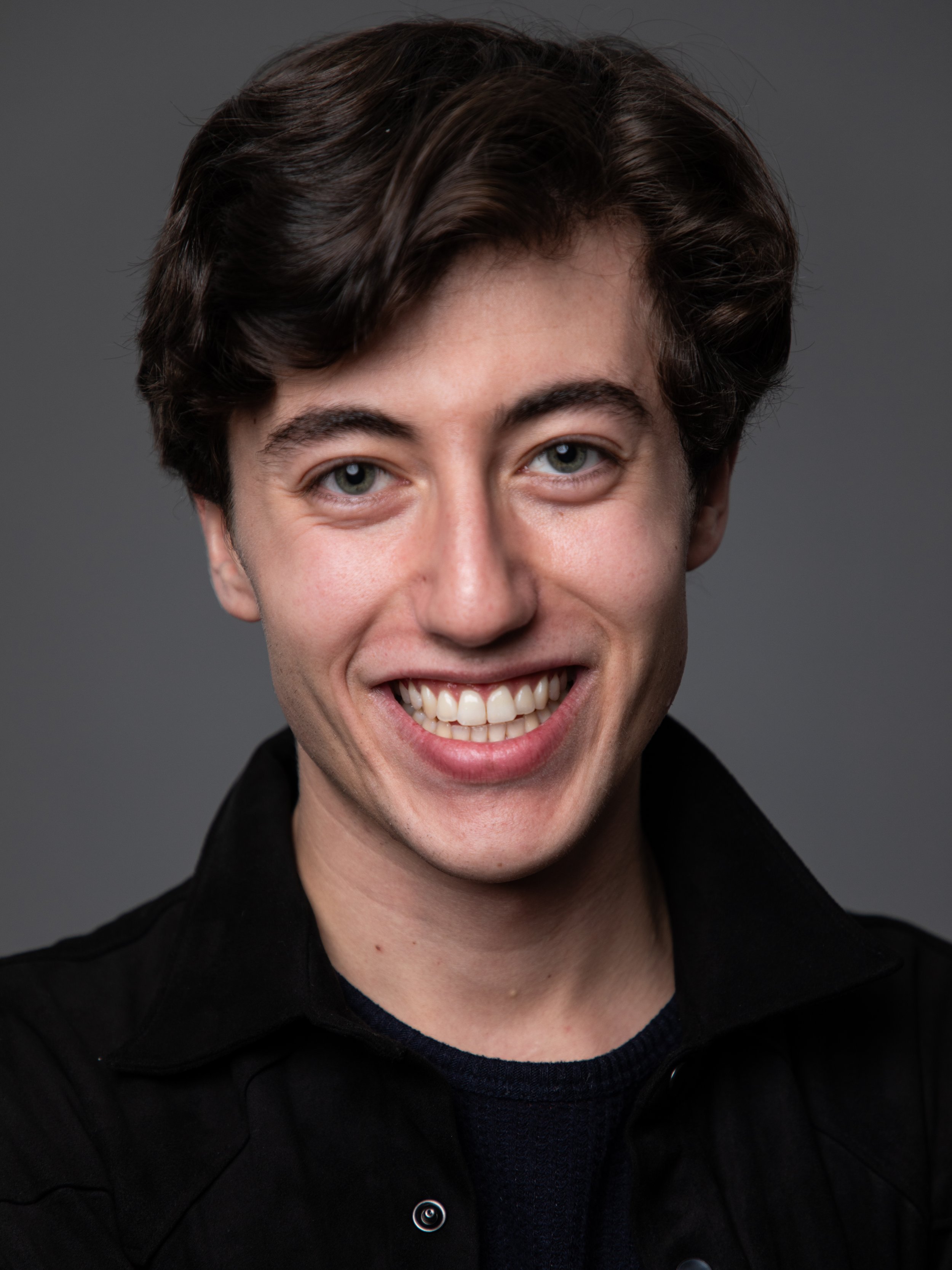 A young man with dark brown hair and blue eyes smiling, wearing a black shirt, against a gray background.