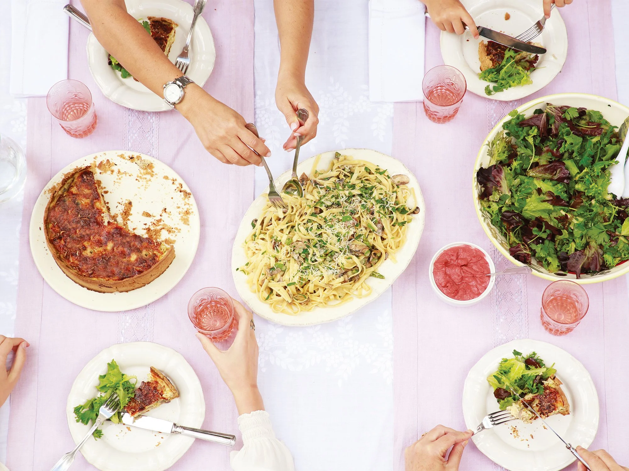 Mesa de jantar com pessoas comendo pizza, massa, salada e bebidas em copos rosa