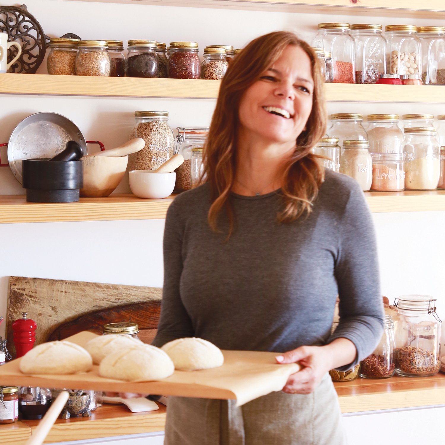 Mulher sorrindo segurando uma tábua com pães frescos, atrás de uma prateleira com potes de ingredientes na cozinha.