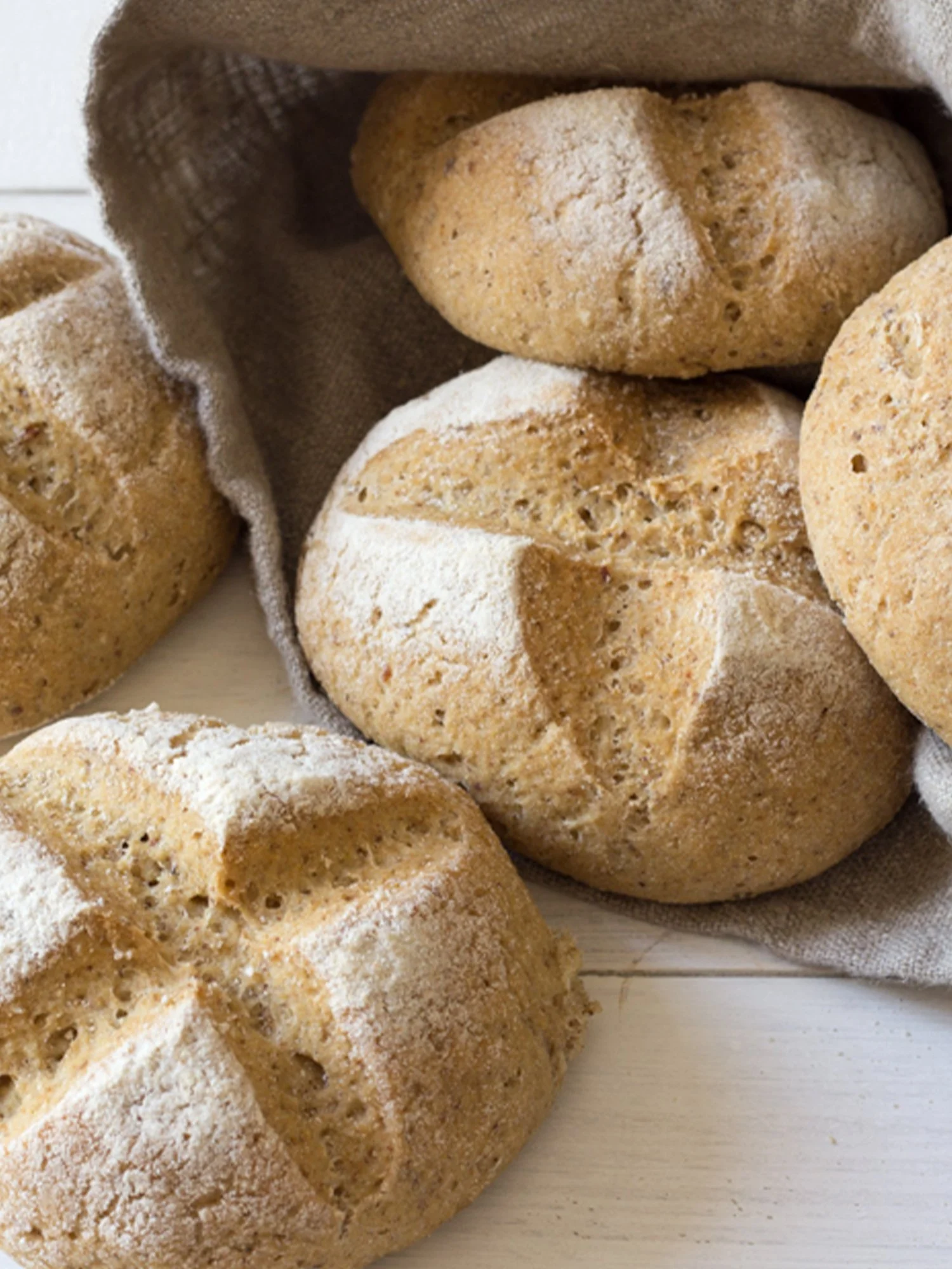 Pãezinhos de pão rústico com farinha espessa, alguns com farinha de trigo polvilhada, em uma toalha de algodão sobre uma superfície de madeira.