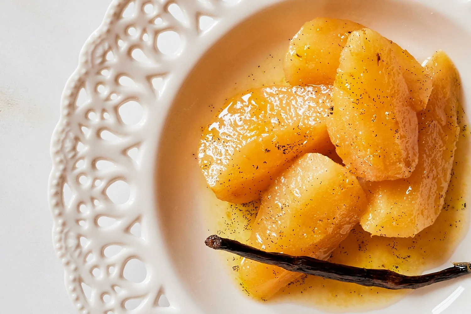 Prato de pera assada com vagem de baunilha e especiarias num prato de louça branca decorada com buracos na borda.