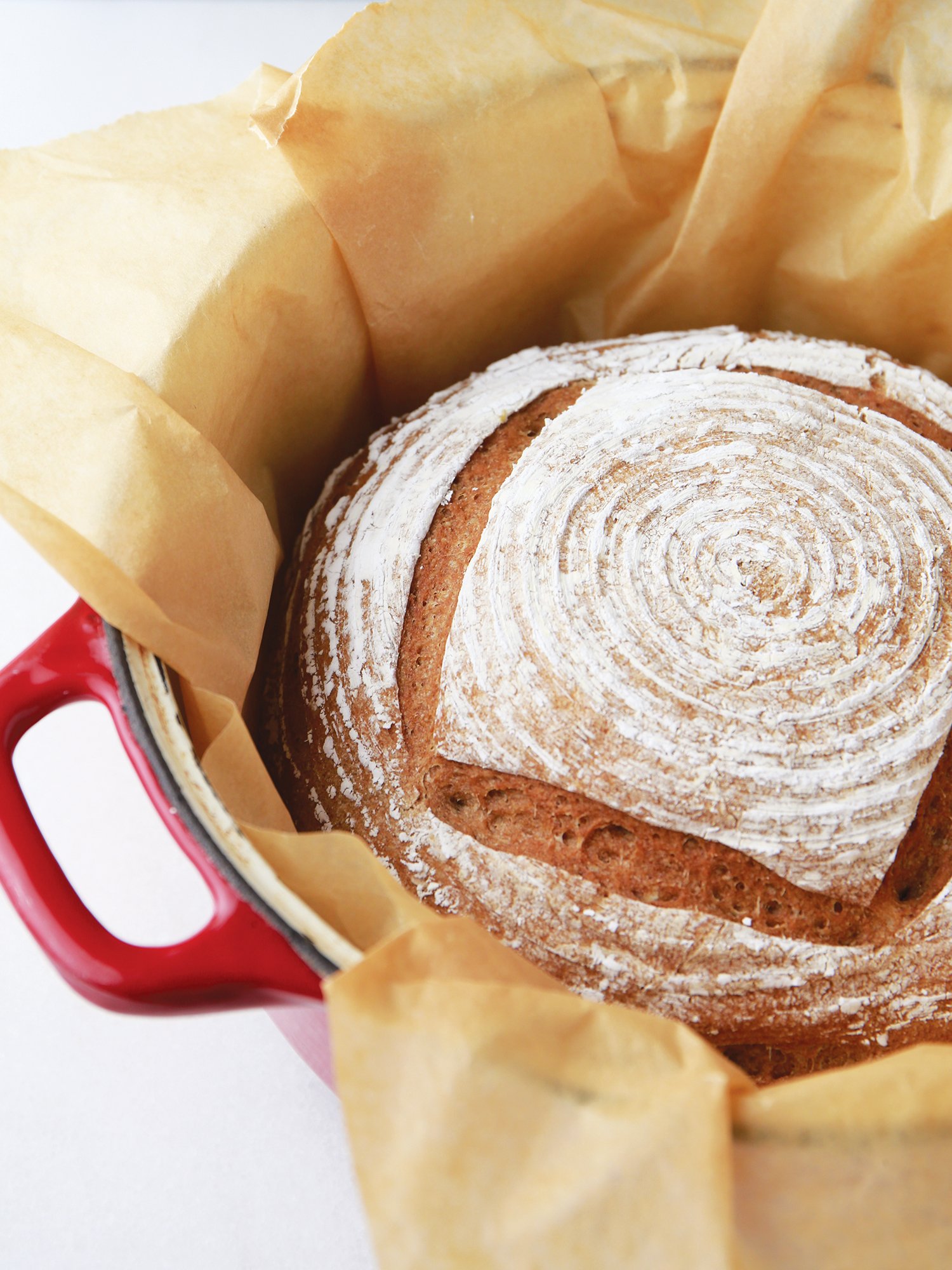 Pão de fermentação natural com crosta avermelhada, polvilhado com farinha, dentro de uma panela de ferro com forro de papel manteiga.