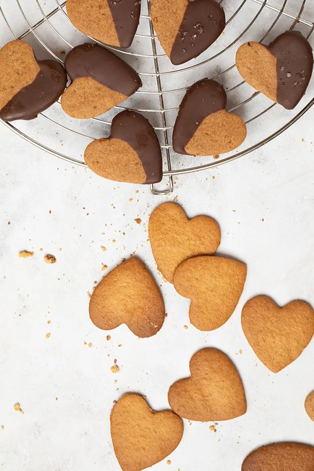 Biscoitos de coração, alguns cobertos com chocolate e outros sem cobertura, sobre uma grade de resfriamento e uma superfície com migalhas de biscoito.