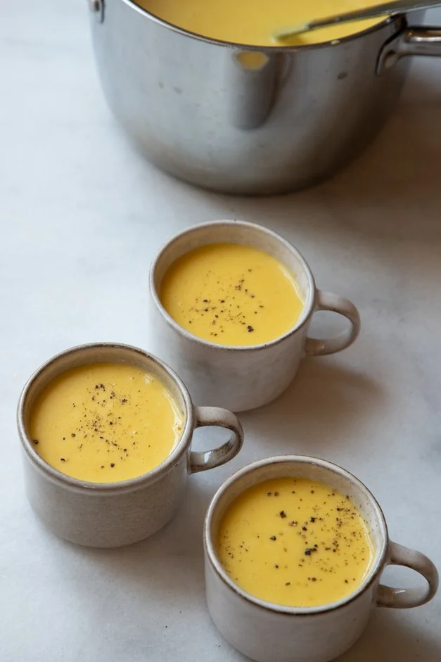 Três xícaras de sopa de milho com pimenta preta, sobre uma mesa branca, ao lado de uma panela de metal com mais sopa de milho.
