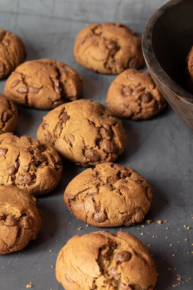 Cookies de chocolate em uma superfície cinza, alguns ao redor de uma tigela de madeira.