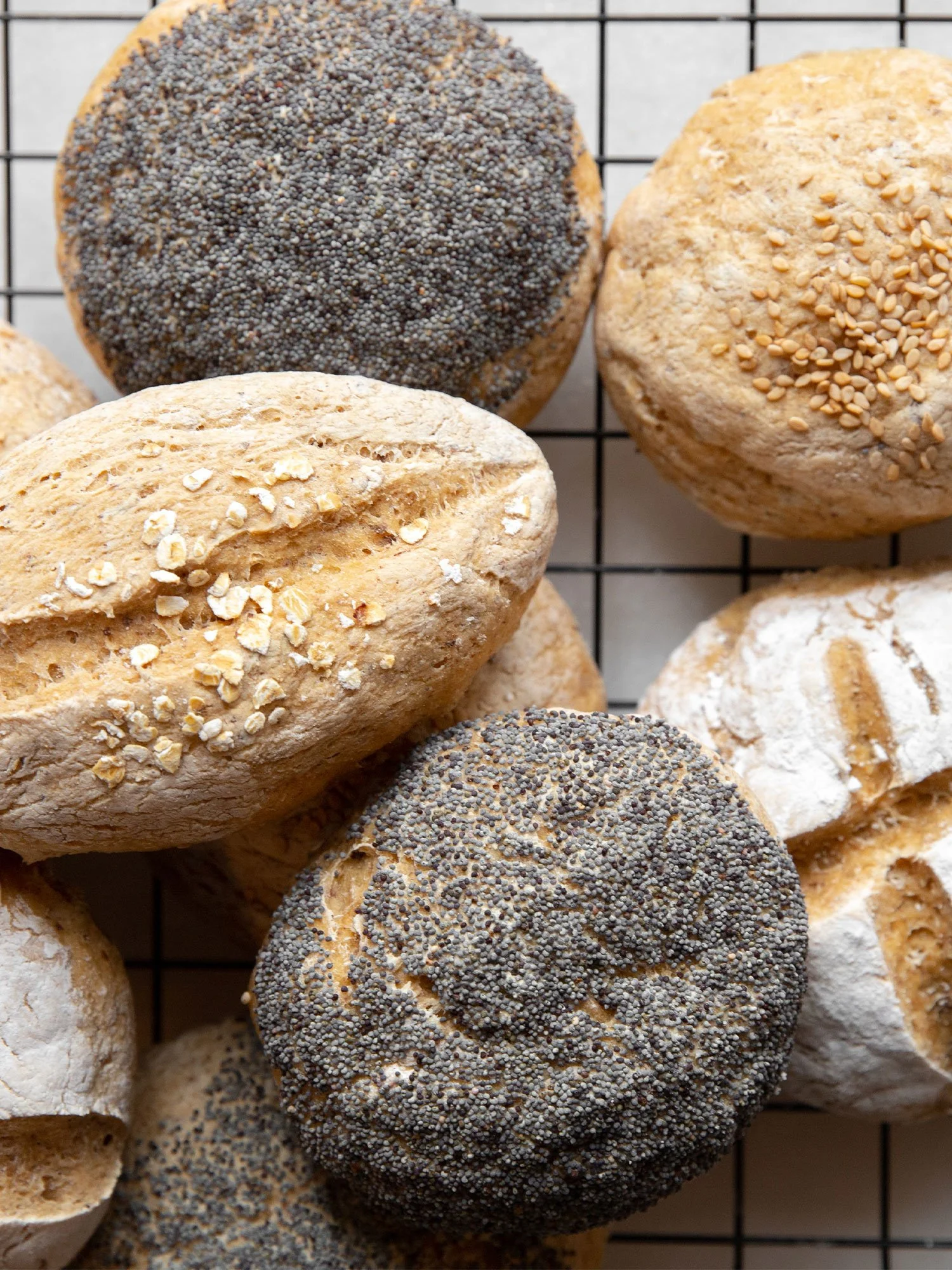 Pãezinhos variados com cobertura de sementes, incluindo gergelim, farinha de trigo e aveia, sobre uma grade de arame.