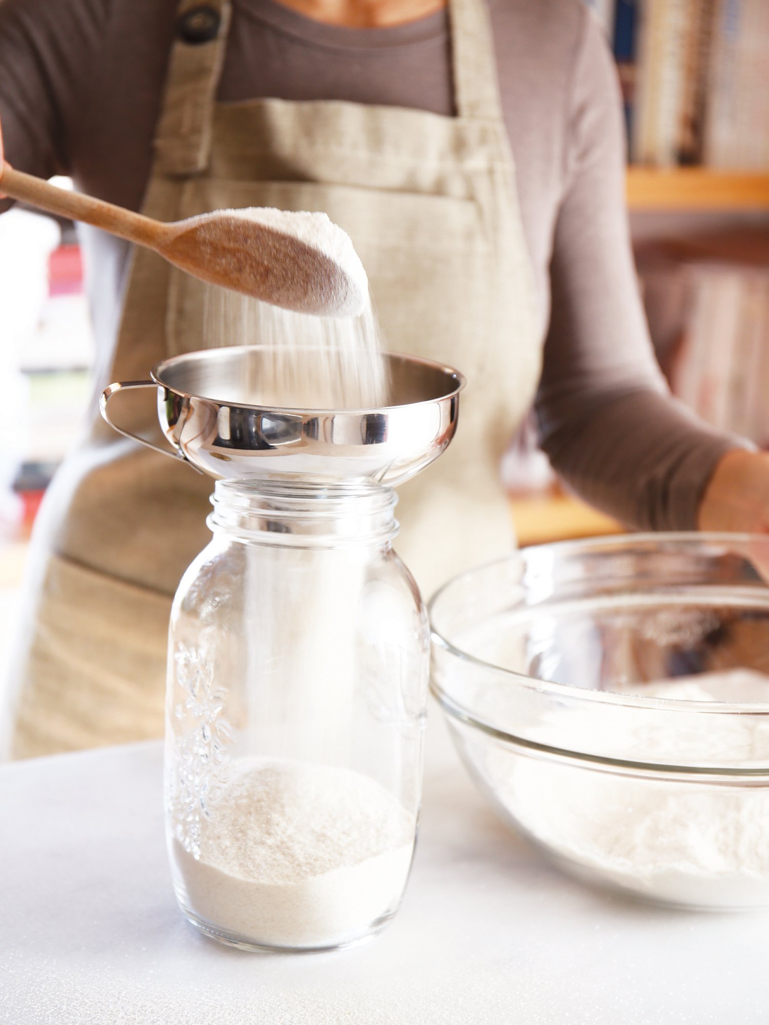 Pessoa colocando farinha em uma peneira sobre um pote de vidro, enquanto outras áreas da cozinha mostram uma tigela com farinha