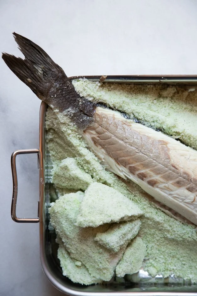 Peixe inteiro com escama, cabeça e cauda, sendo preparado para assar, coberto com farinha de pão, dentro de uma assadeira de metal.