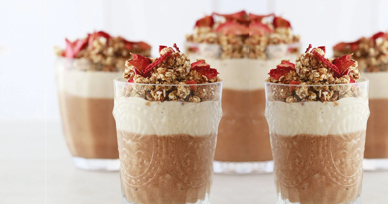 Copa de sobremesa com camadas de mousse de chocolate, chantilly e granola com pedaços de frutas vermelhas por cima, com um bolo de chocolate ao fundo.
