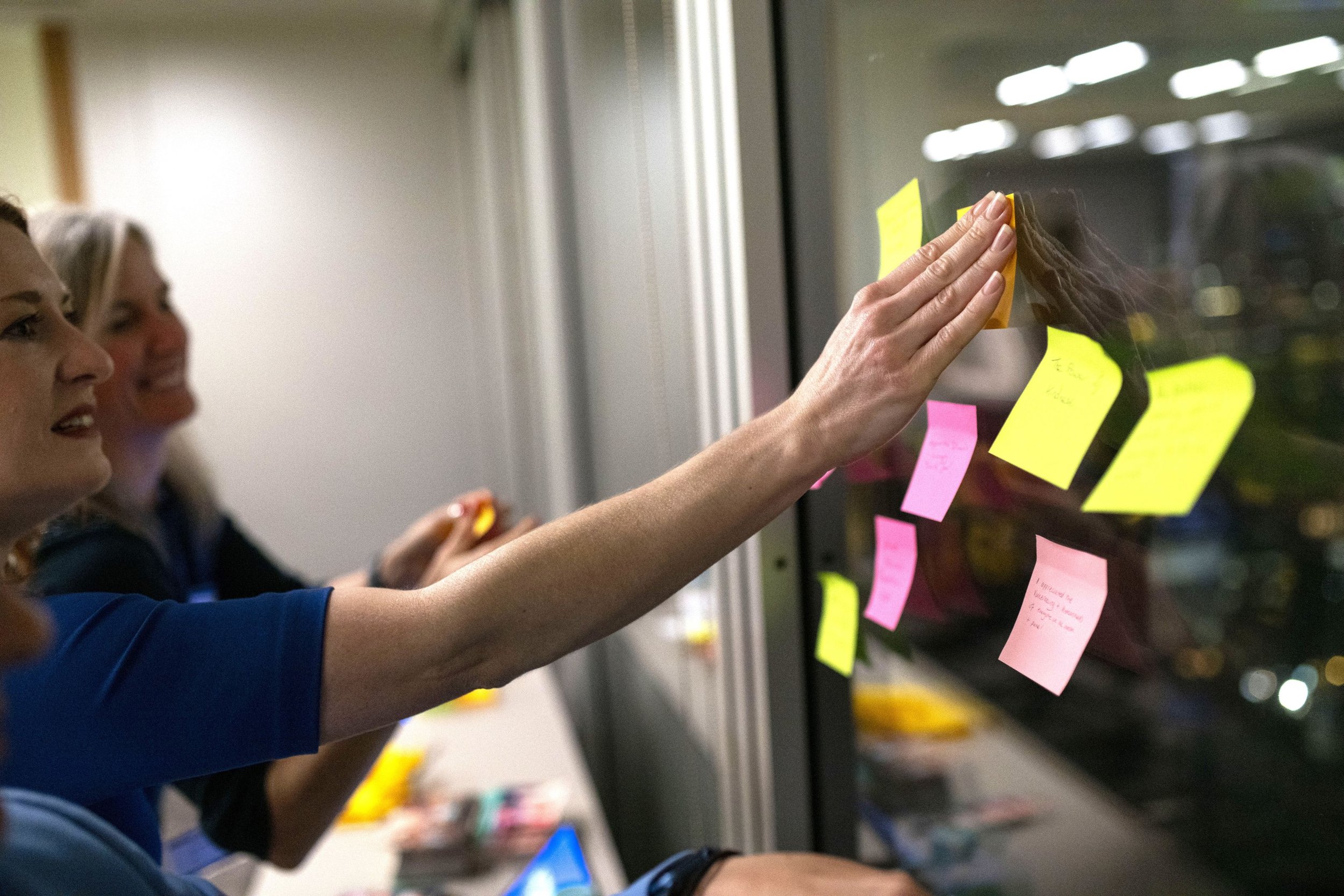 A profile view of Diana Squires consulting on a London business project. She is smiling and attaching post-its to a window, showing the creative research and development work she completed for the National Centre for Academic and Cultural Exchange.