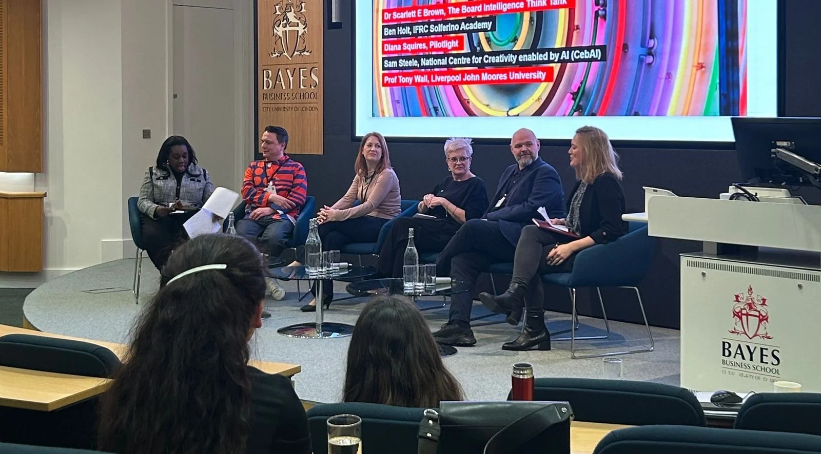 Diana Squires onstage with 4 other professional panelists, speaking at World Creativity & Innovation Day in London. Behind them is a red Bayes Business School plaque and presentation screen.