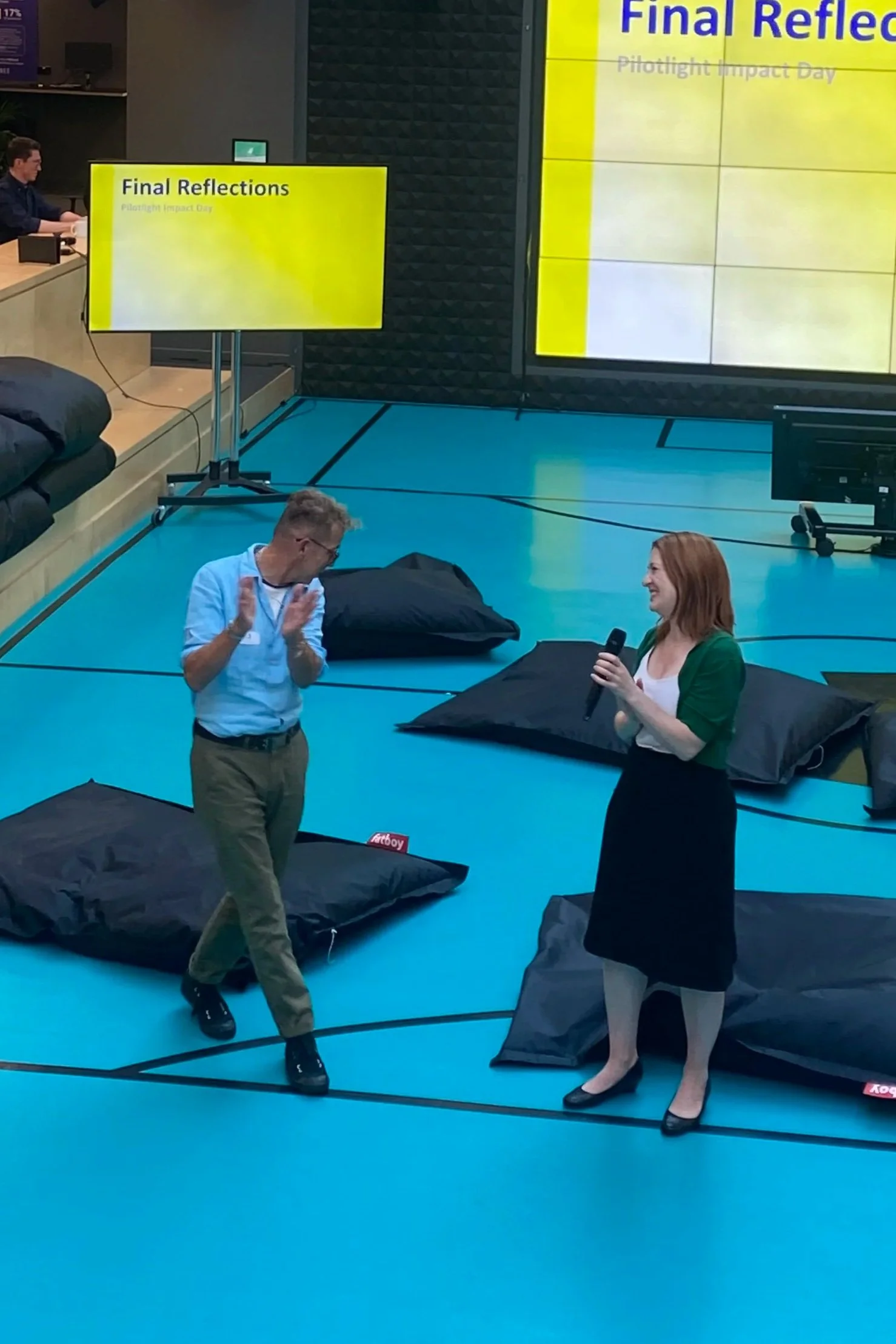 Inspirational speaker Diana Squires and a male charity leader speak into handheld mics in an innovation hub surrounded by bean-bag chairs and yellow presenting screens.