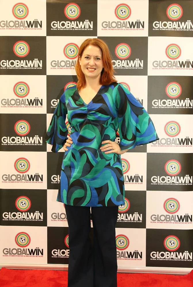 London workshop presenter Diana Squires stands with hands on her hips in a flowing teal shirt, with the GlobalWIIN conference banner behind her.