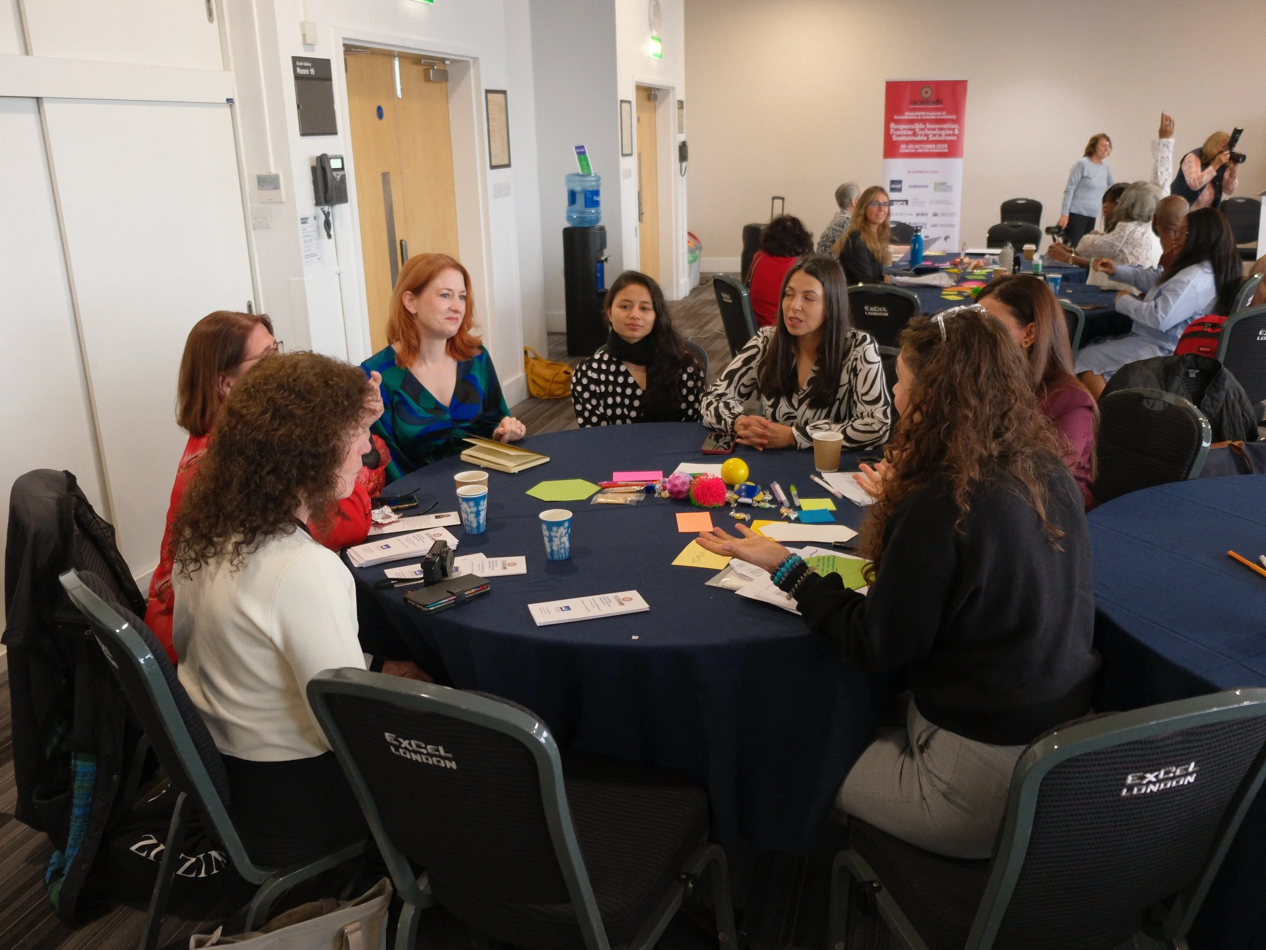 Diana Squires MSc facilitates a table conversation at a social innovation event. 6 female entrepreneurs discuss strategy with her. Colourful ideation tools and gadgets are clustered in the middle of the table. Behind, a banner saying GlobalWIIN.