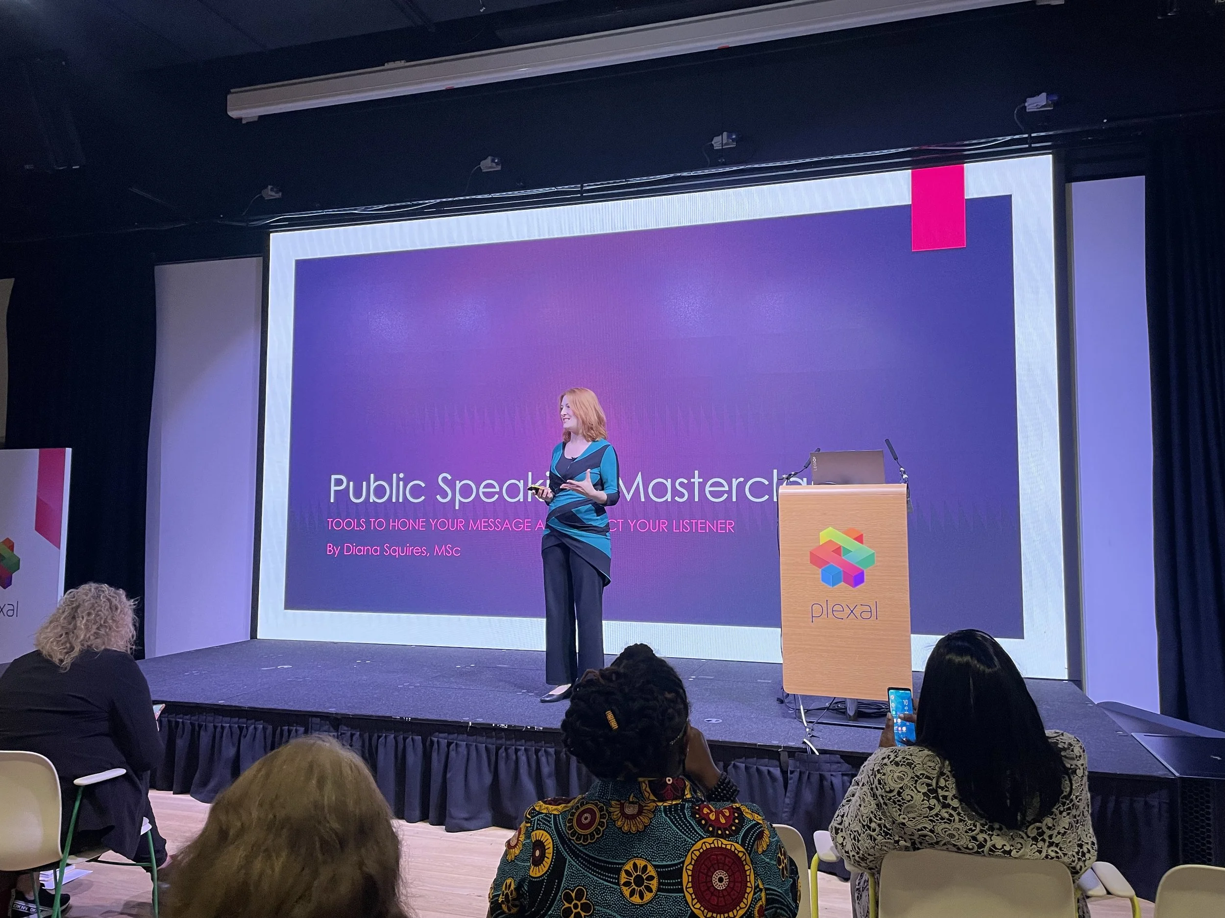 Workshop presenter Diana Squires stands confidently on a stage as a featured speaker at a conference for the Global Women Inventors & Innovators Network. The screen says: Public SpeakingMasterclass, Tools to Hone Your Message and Impact Your Speaker.