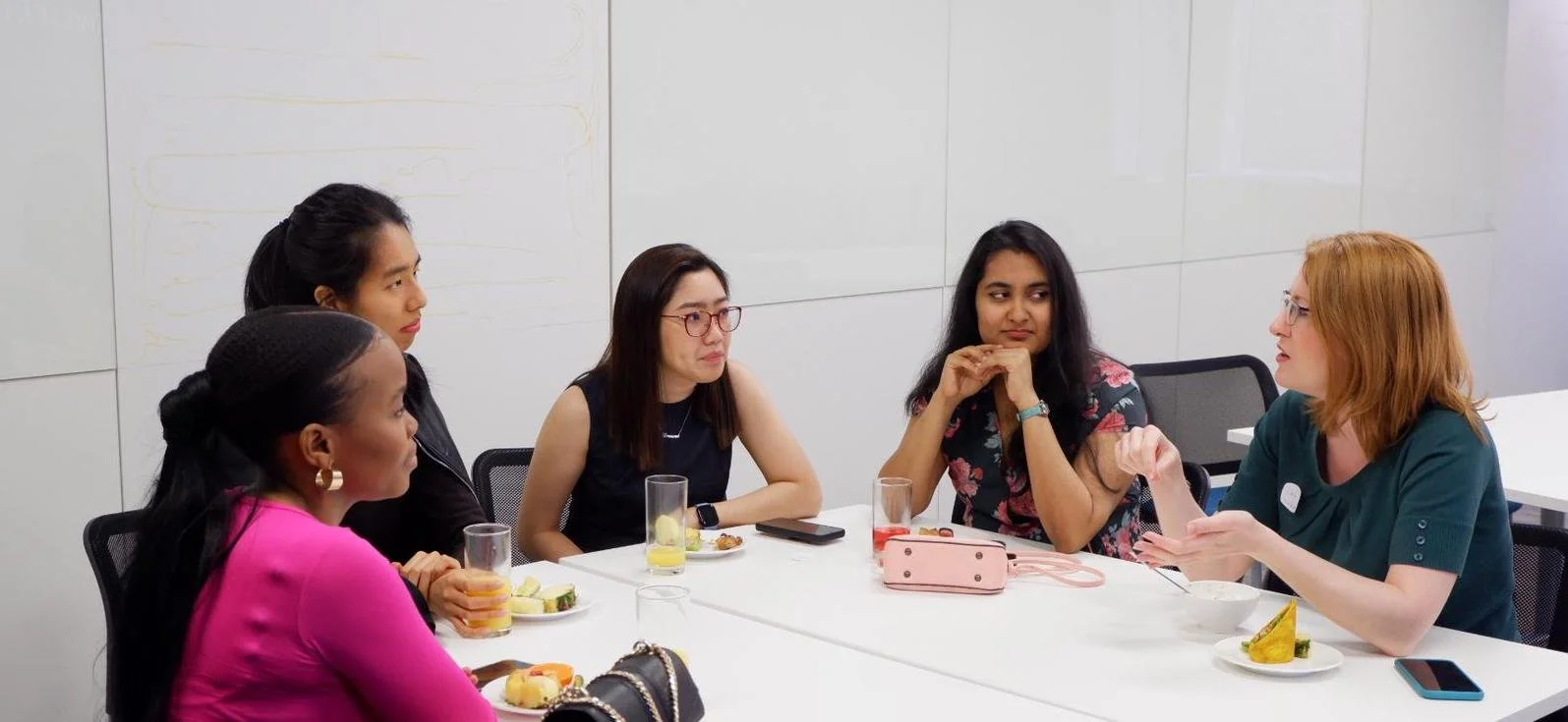 In a corporate meeting room, a mentorship group of four women sit at a table listening to the mentor and facilitator, Diana Squires. She sits on the right, wearing a business-casual shirt and using her hands to explain a concept.