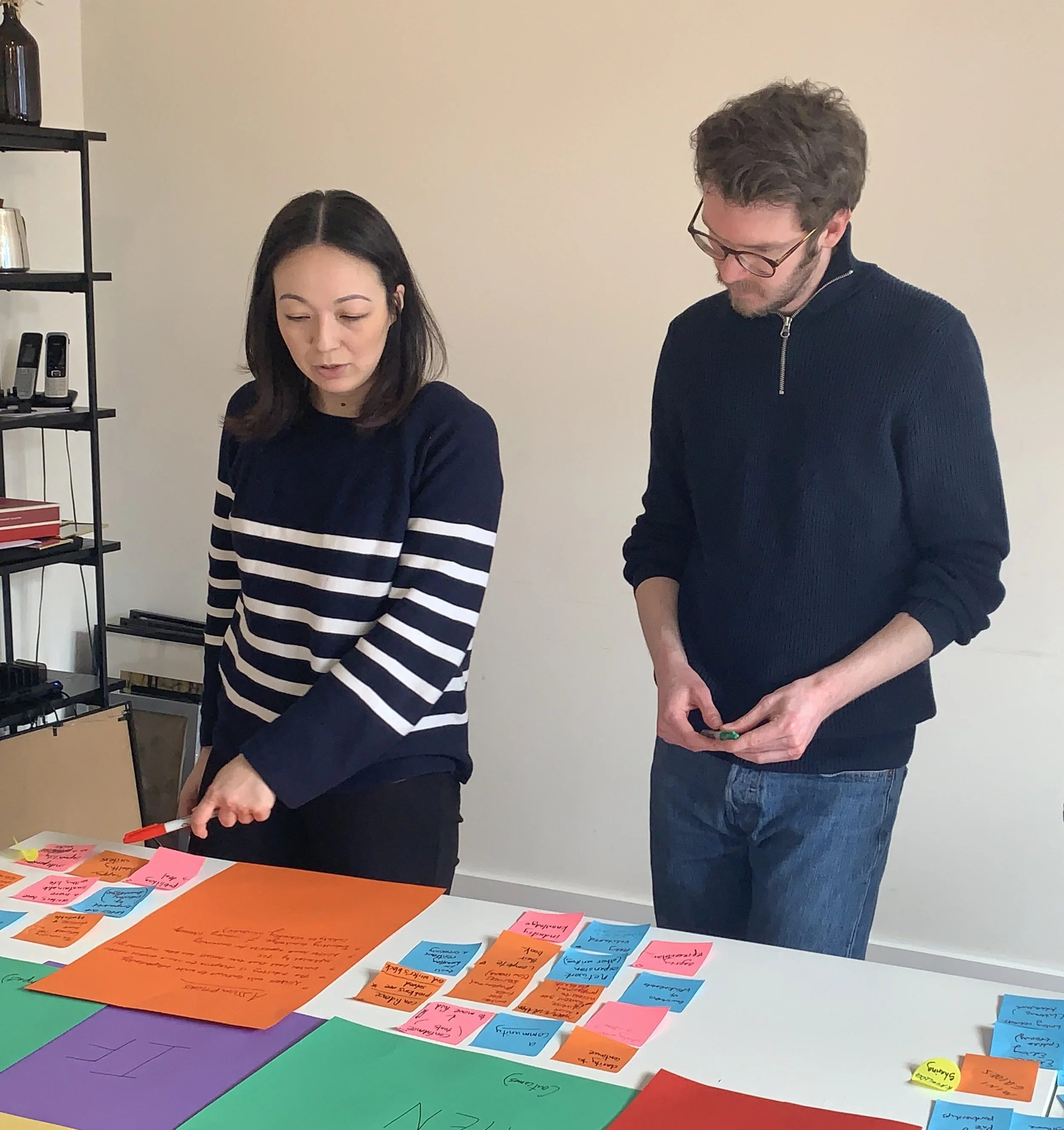 An Asian woman wearing a striped jumper and a younger caucasian male wearing a zip-sweater engage with post-its spread out on a table in a strategy mapping session