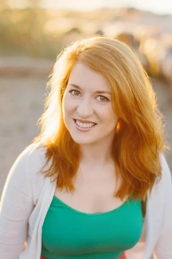 Consulting headshot of a young Diana Squires smiling at the camera. She has long red hair and the sun illuminating her from behind. She wears a bright green shirt with a white cardigan.