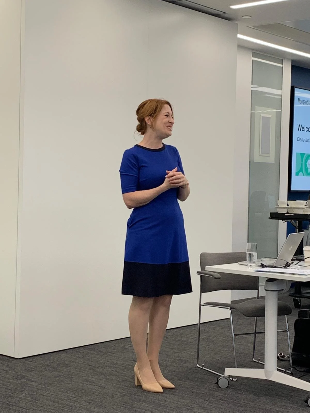Strategic consultant Diana Squires stands at a whiteboard with a speaker's table next to her, addressing a group of people (out of frame) at a charity consulting programme. She wears a cheerful expression, red hair, blue dress and heels.