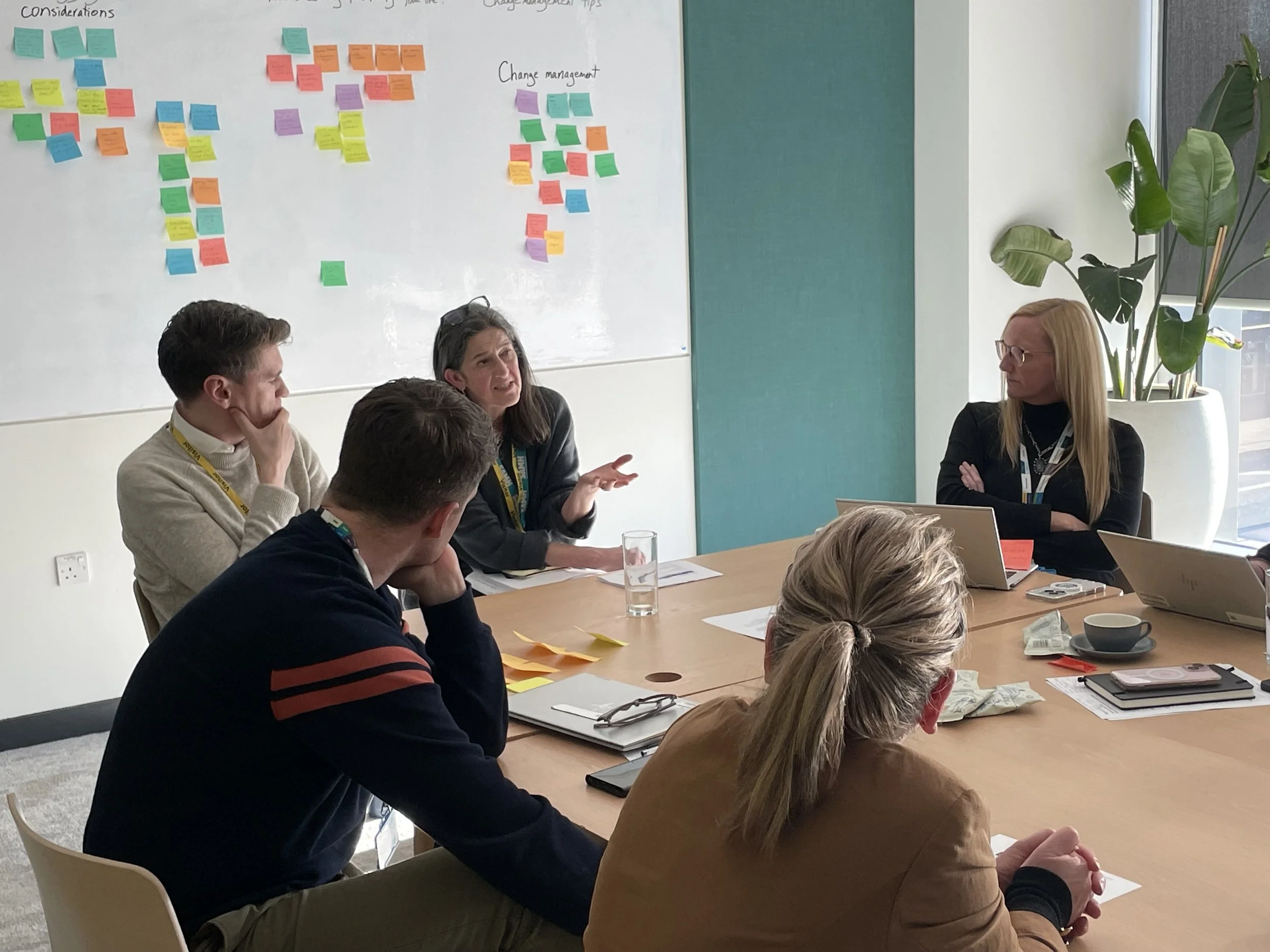 At an idea generation event (UK) a female charity consultant with dark hair explains an organisational resilience challenge to a team that Diana coached and facilitated. Behind their collaboration table is a design thinking whiteboard with post-its.