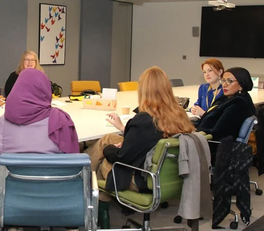 5 women sit around a table talking, three facing the camera, in a business setting. They are ethnically diverse and one is consultant Diana Squires.