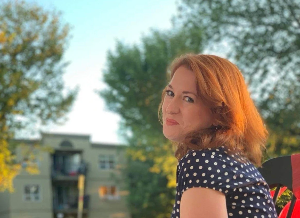 Casual headshot of speaker and facilitator Diana Squires, sitting outdoors in summer and smiling sideways at the camera. She wears a navy, polka-dot dress and has curls in her long red hair. A tree is blurred behind her.