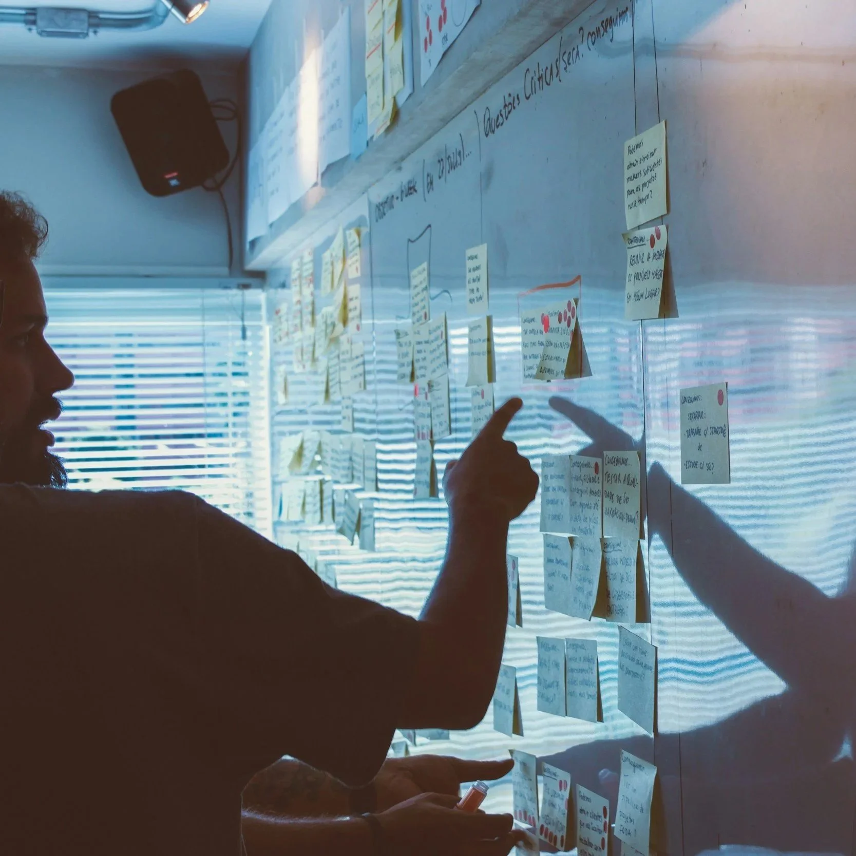 Person pointing at notes on a whiteboard with sticky notes and handwritten text, sunlight coming through blinds in the background.