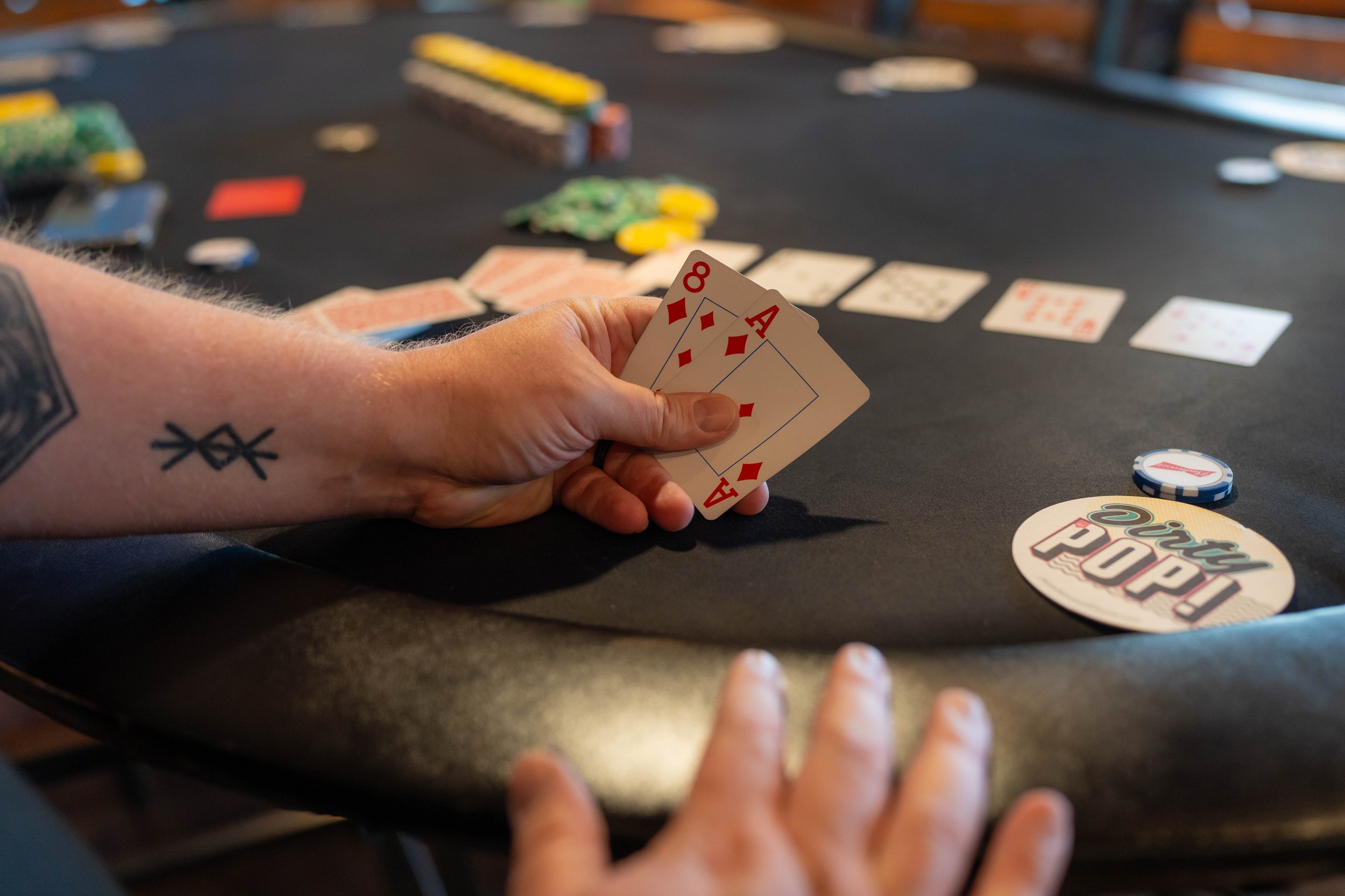 A person holding four playing cards, including the 8 and Ace of diamonds, during a game of poker at a black felt table, with poker chips and a dealer button nearby.