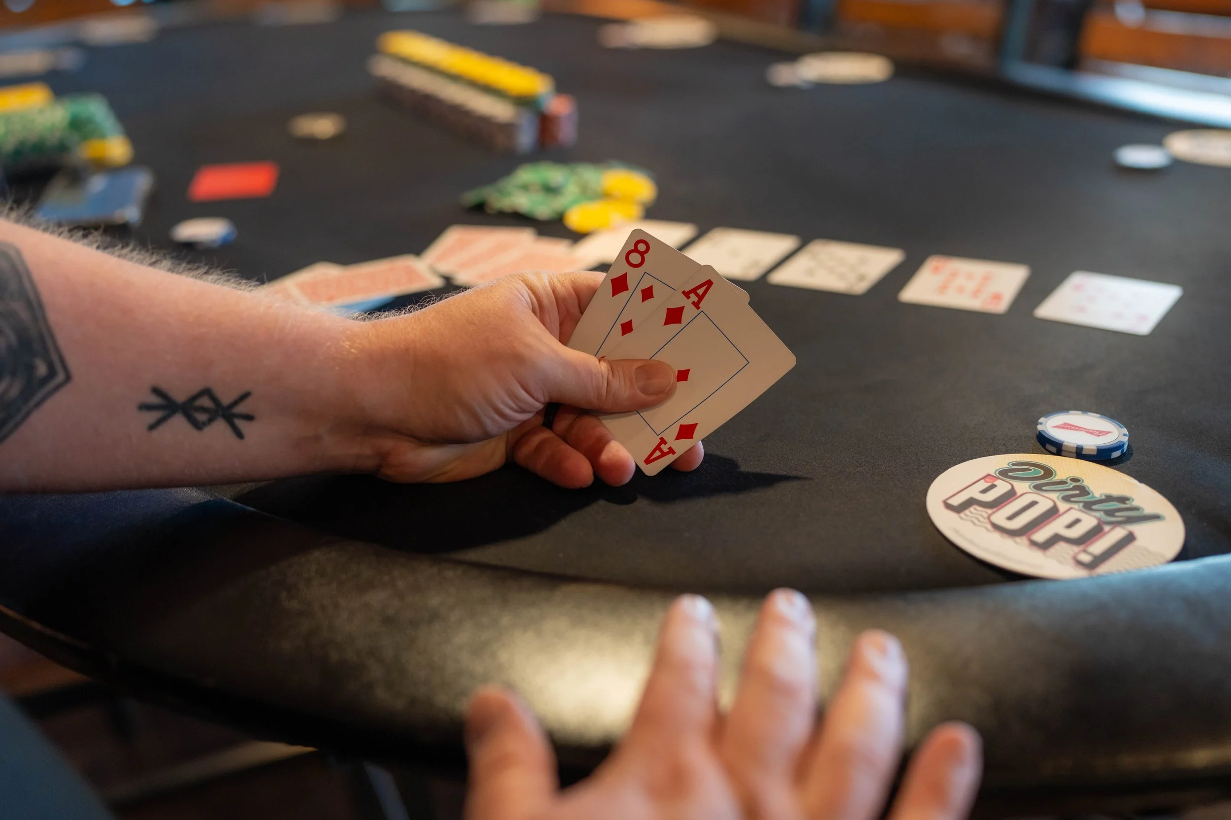 A person holding four playing cards, including the 8 and Ace of diamonds, during a game of poker at a black felt table, with poker chips and