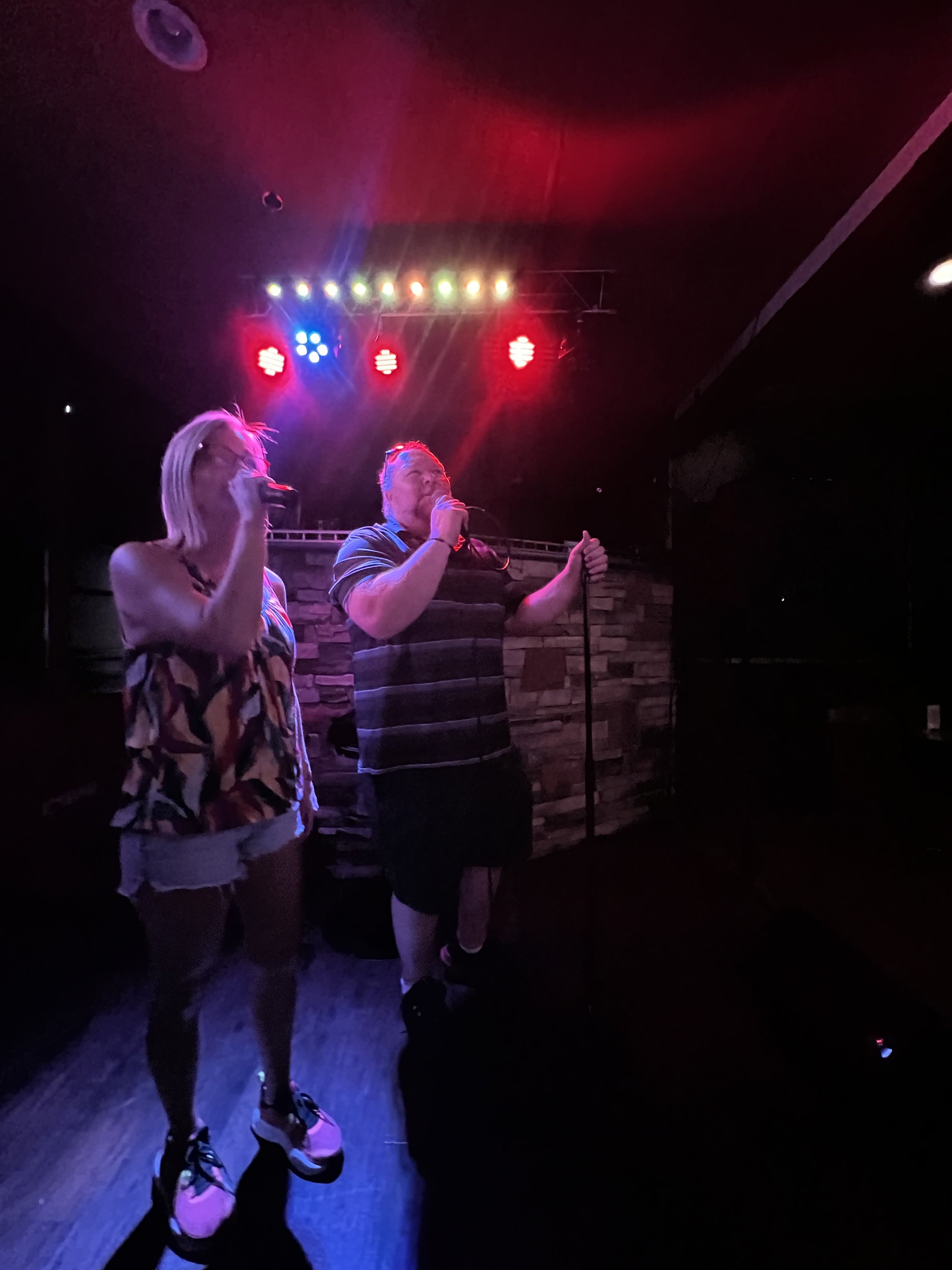 Two people singing into microphones on a stage with colorful lights and a brick wall background.