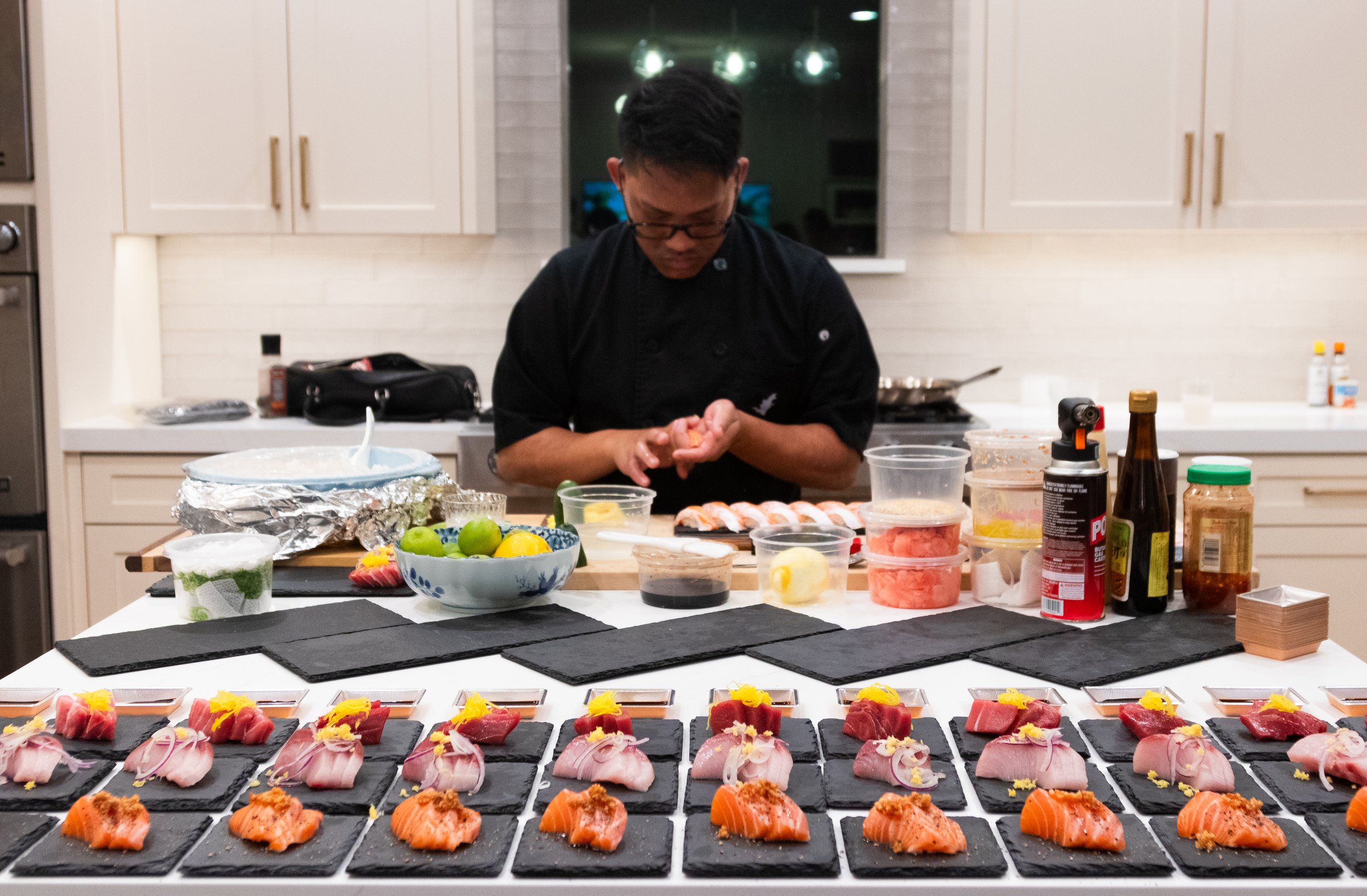 A chef preparing sushi on a kitchen countertop with various ingredients and dishes, with multiple plates of sashimi and sushi ready for serving in front.