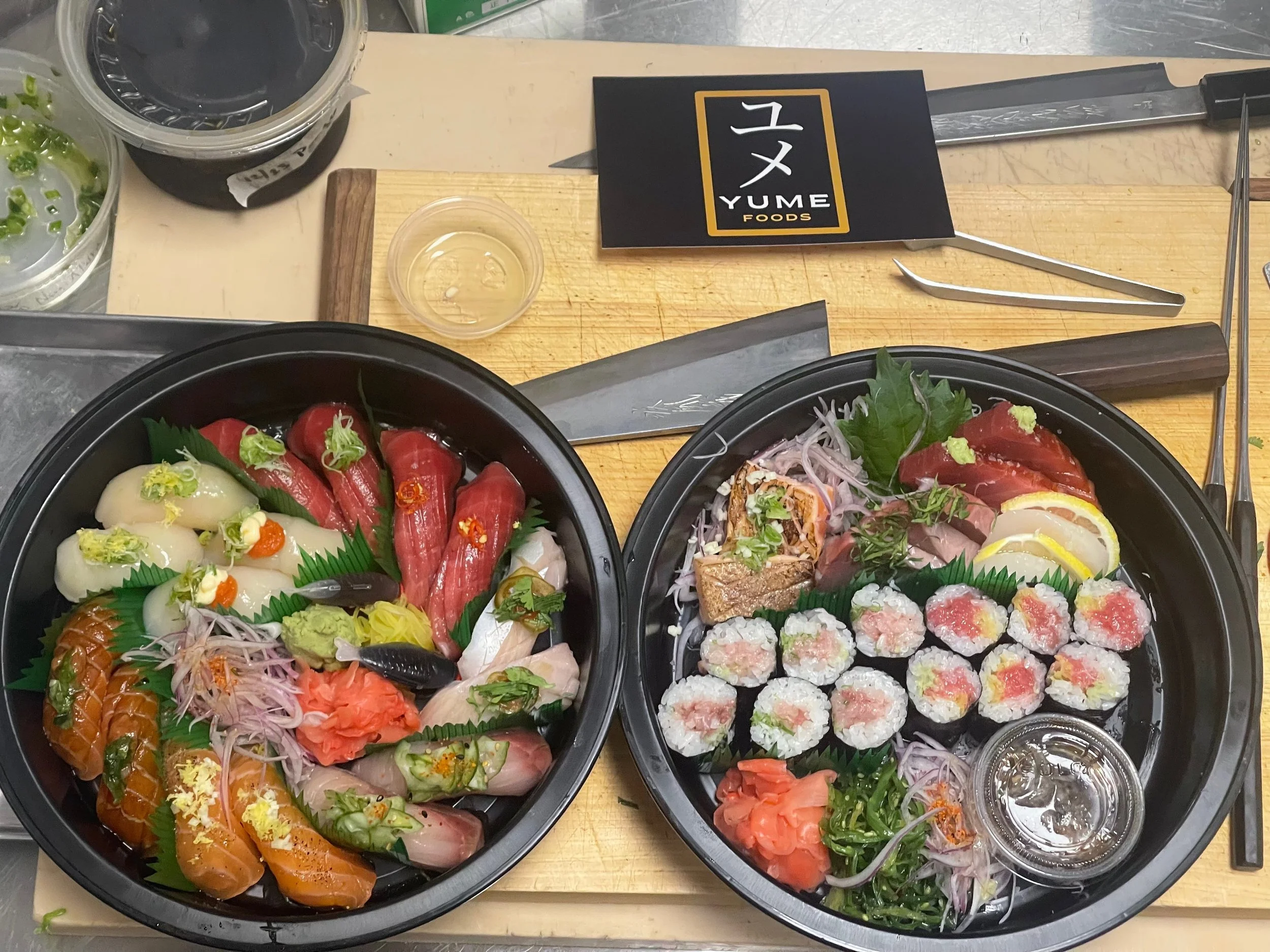 Two black sushi platters with assorted sushi, sashimi, and garnishes on a wooden cutting board. One platter includes salmon, tuna, and white fish sashimi, and sliced rolls, while the other features various sashimi, sliced rolls, and garnishes like wasabi, pickled ginger, and seaweed. A small cup of soy sauce and a small container of water are also visible.