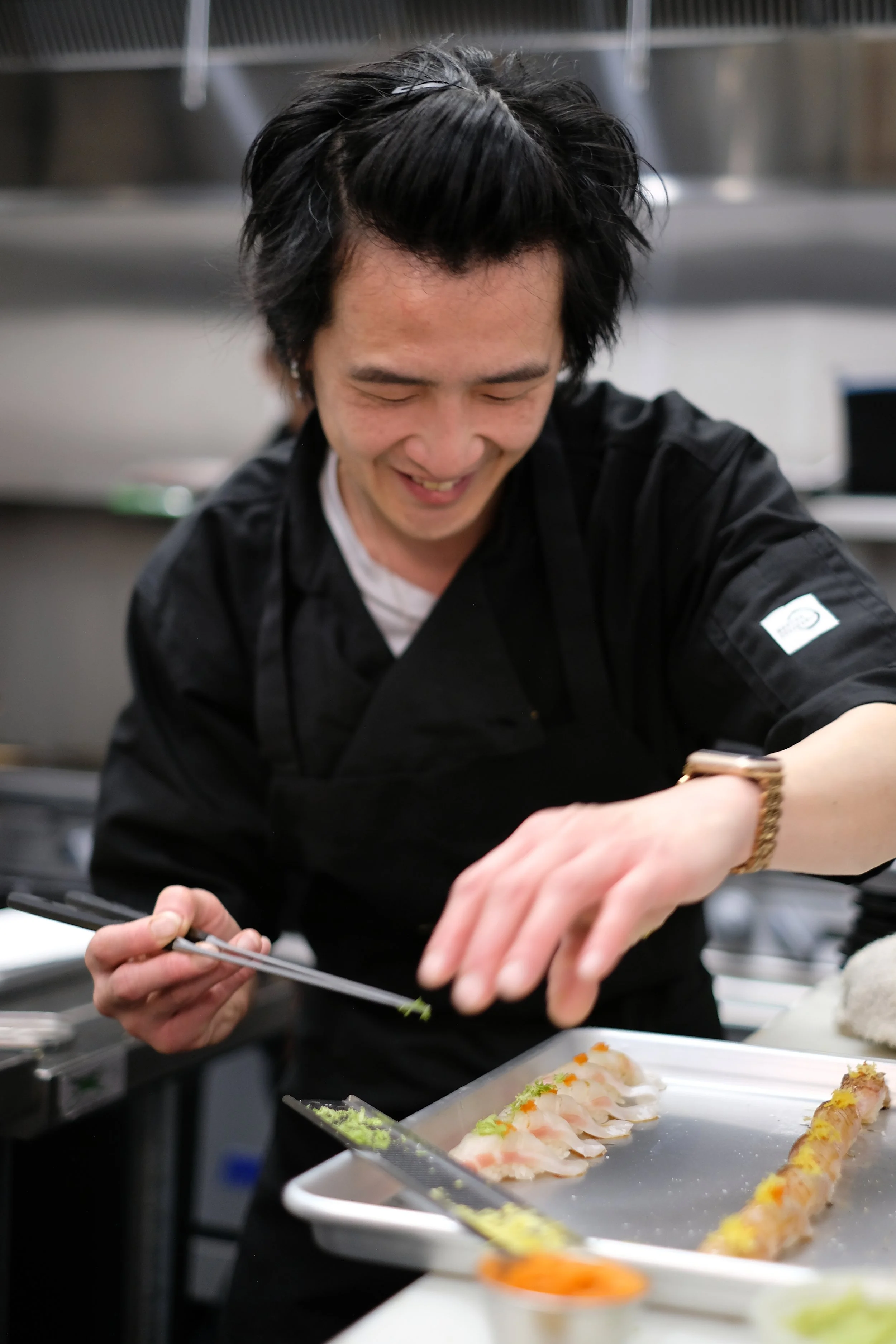 A chef in black attire preparing sushi in a professional kitchen, smiling while garnishing a plate of sliced fish.