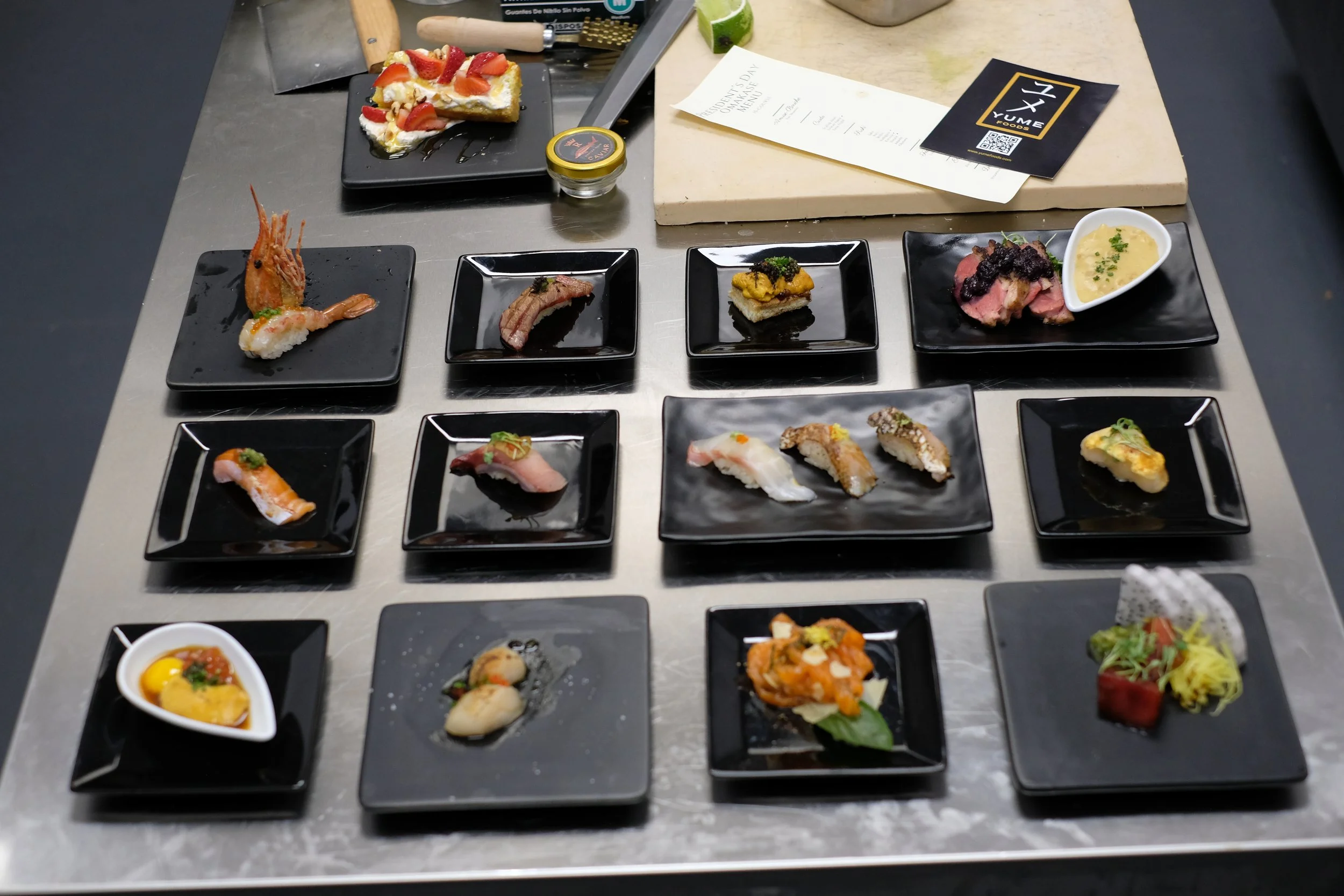 Array of assorted sushi dishes on black plates, with a slice of cake and a menu in the background.