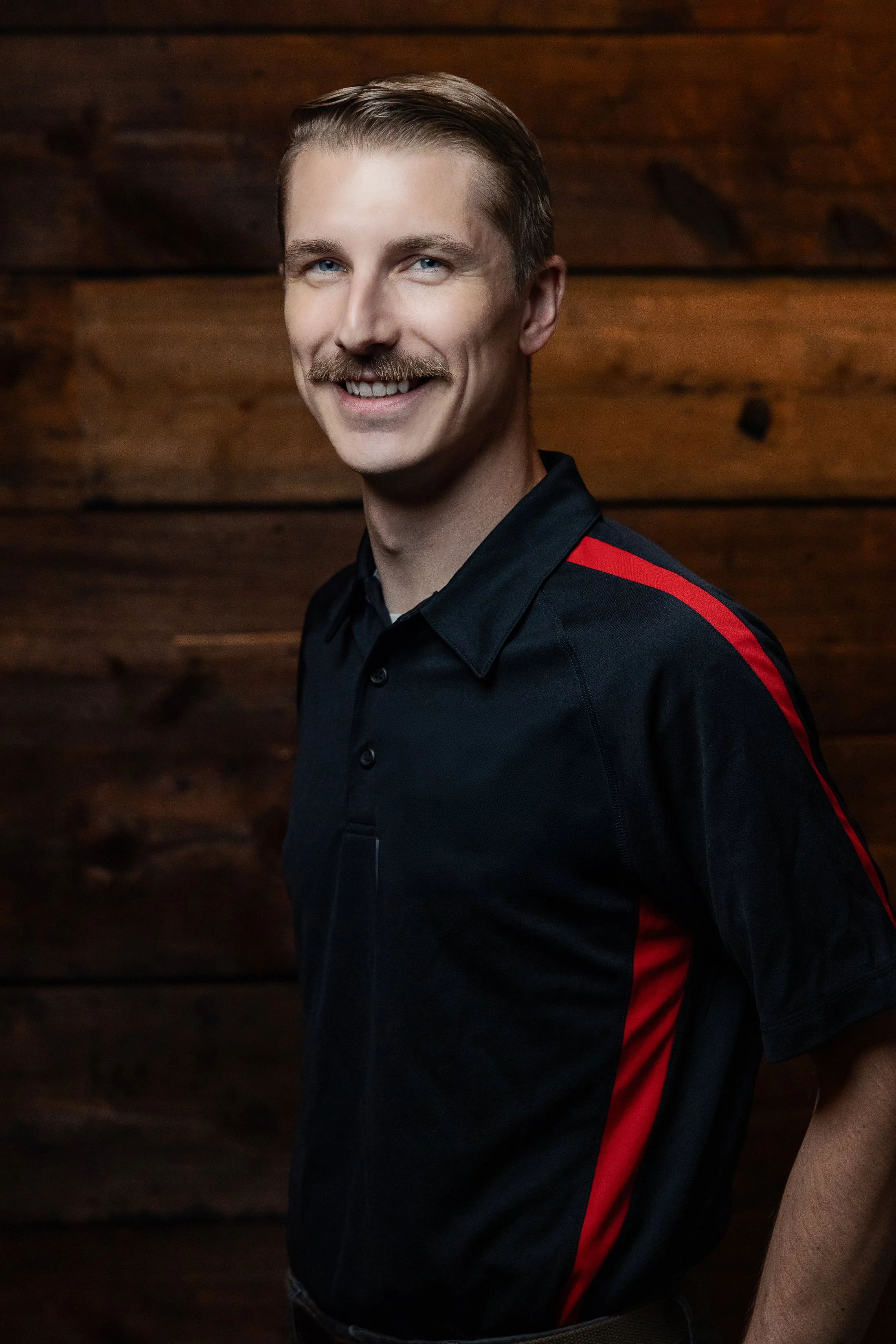 Founder portrait of Will Harkness. A  man with light skin, light brown hair styled back, blue eyes, a mustache, and a friendly smile, wearing a black shirt, standing against a dark wooden background.