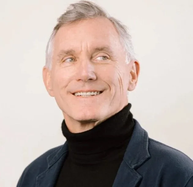 A smiling man with gray hair wearing a black turtleneck and a dark blazer, looking to the side against a plain white background.